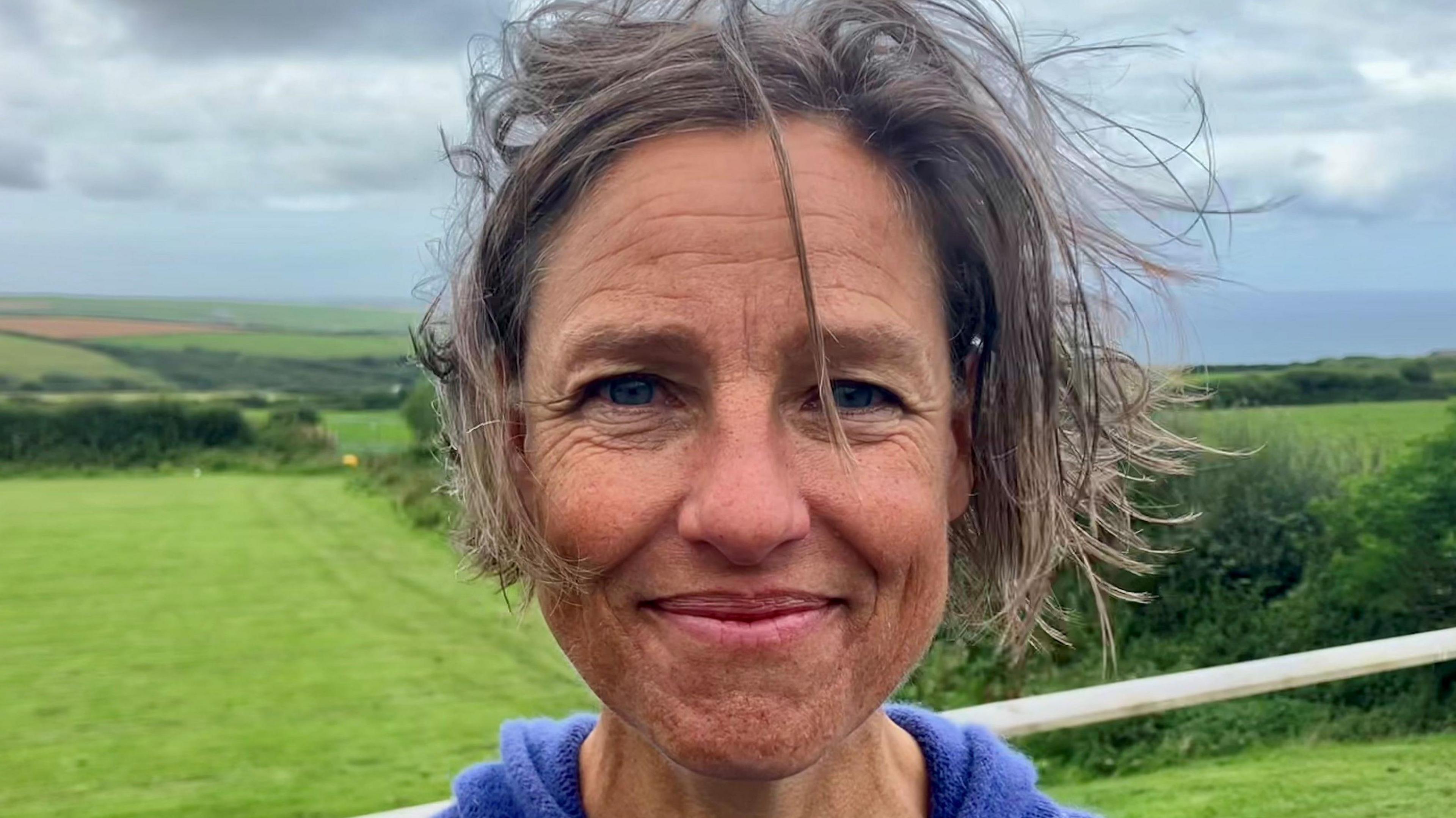 Liz George, with short brown hair, smiling standing in front of fields, with Port Issac Bay in the distance 