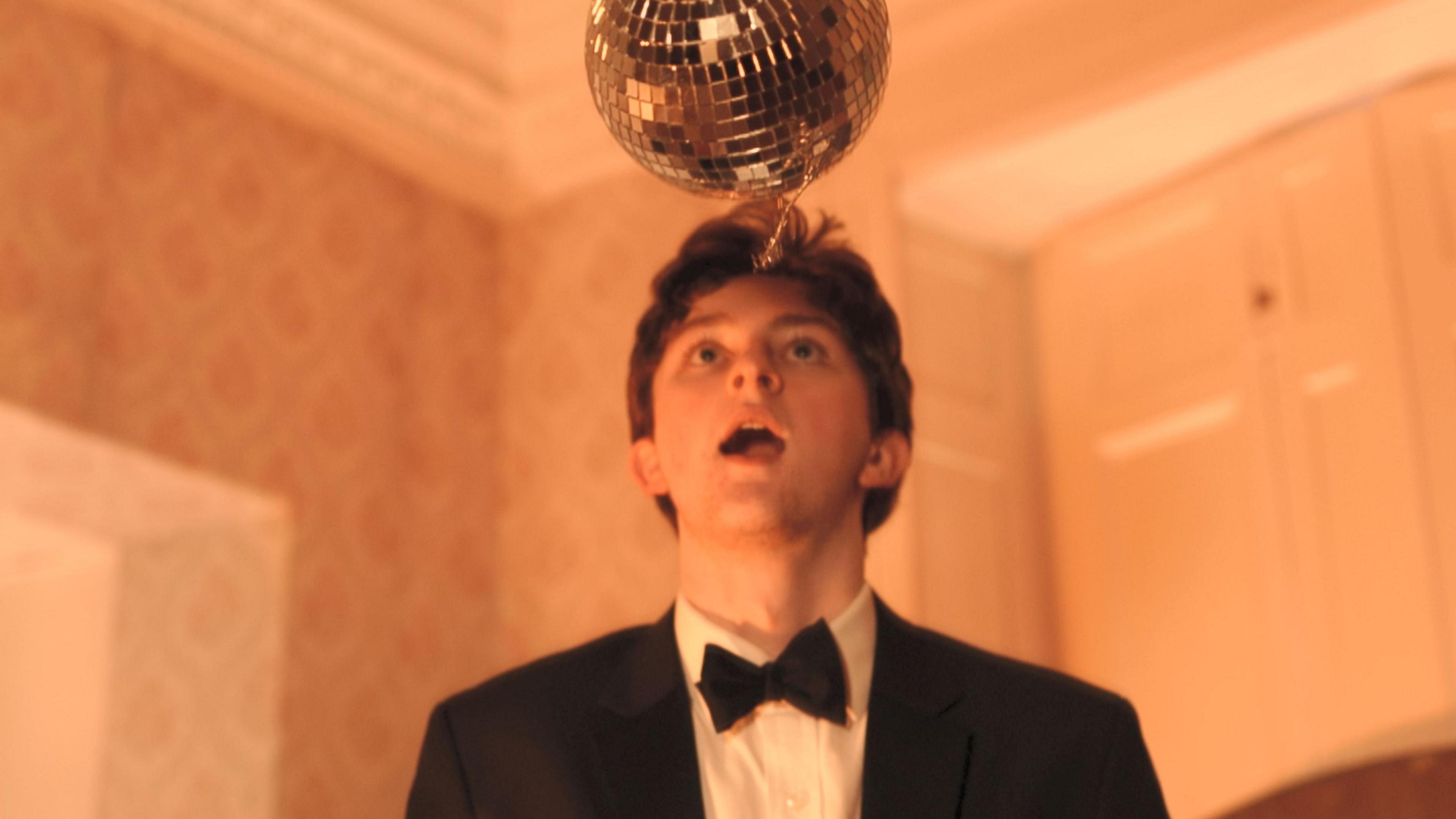 A man is wearing a black suit with a bowtie. He is looking up at a disco ball. 