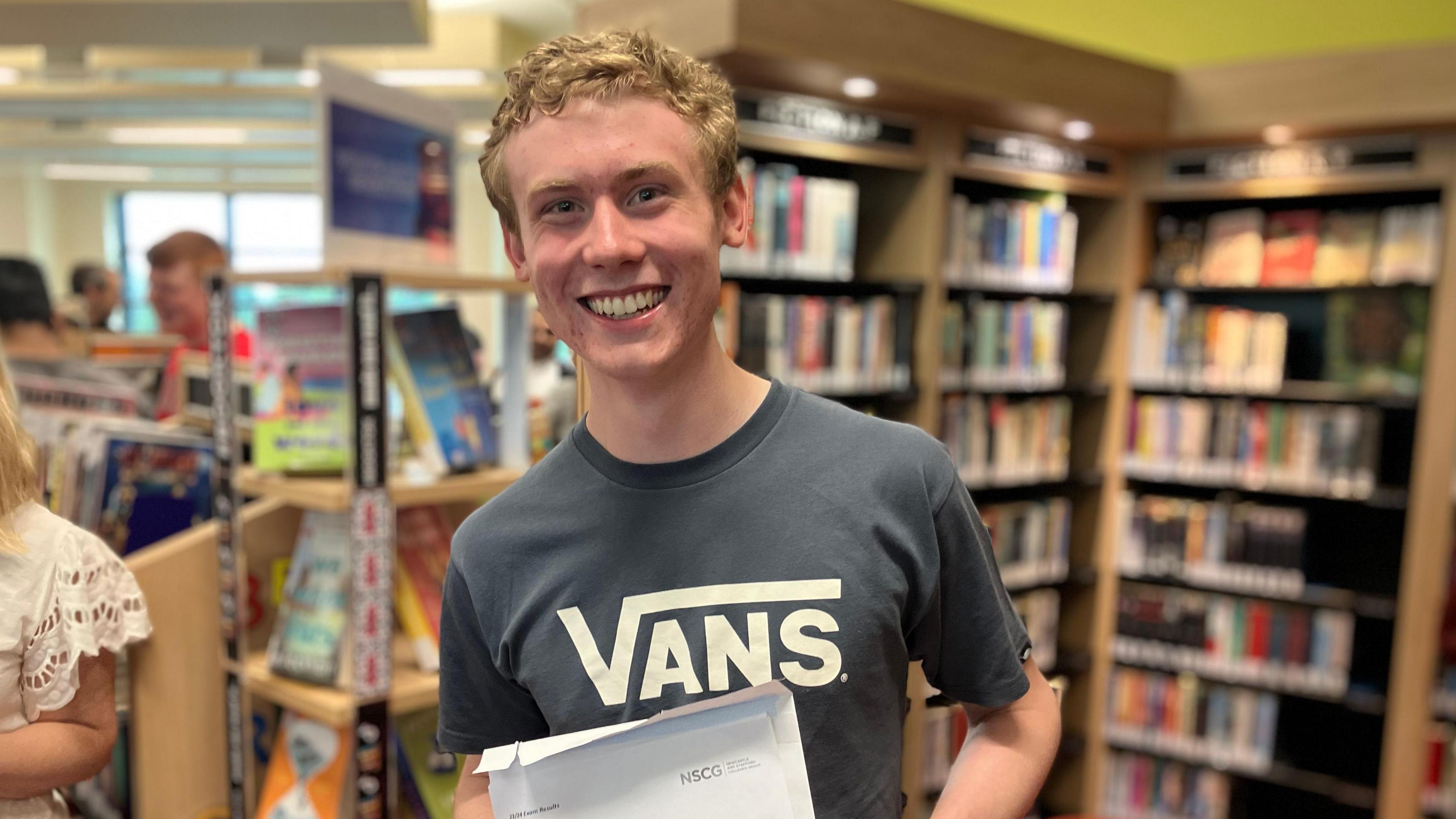 A man stands smiling while holding a paper sheet containing his A-level results with a bookcase behind him