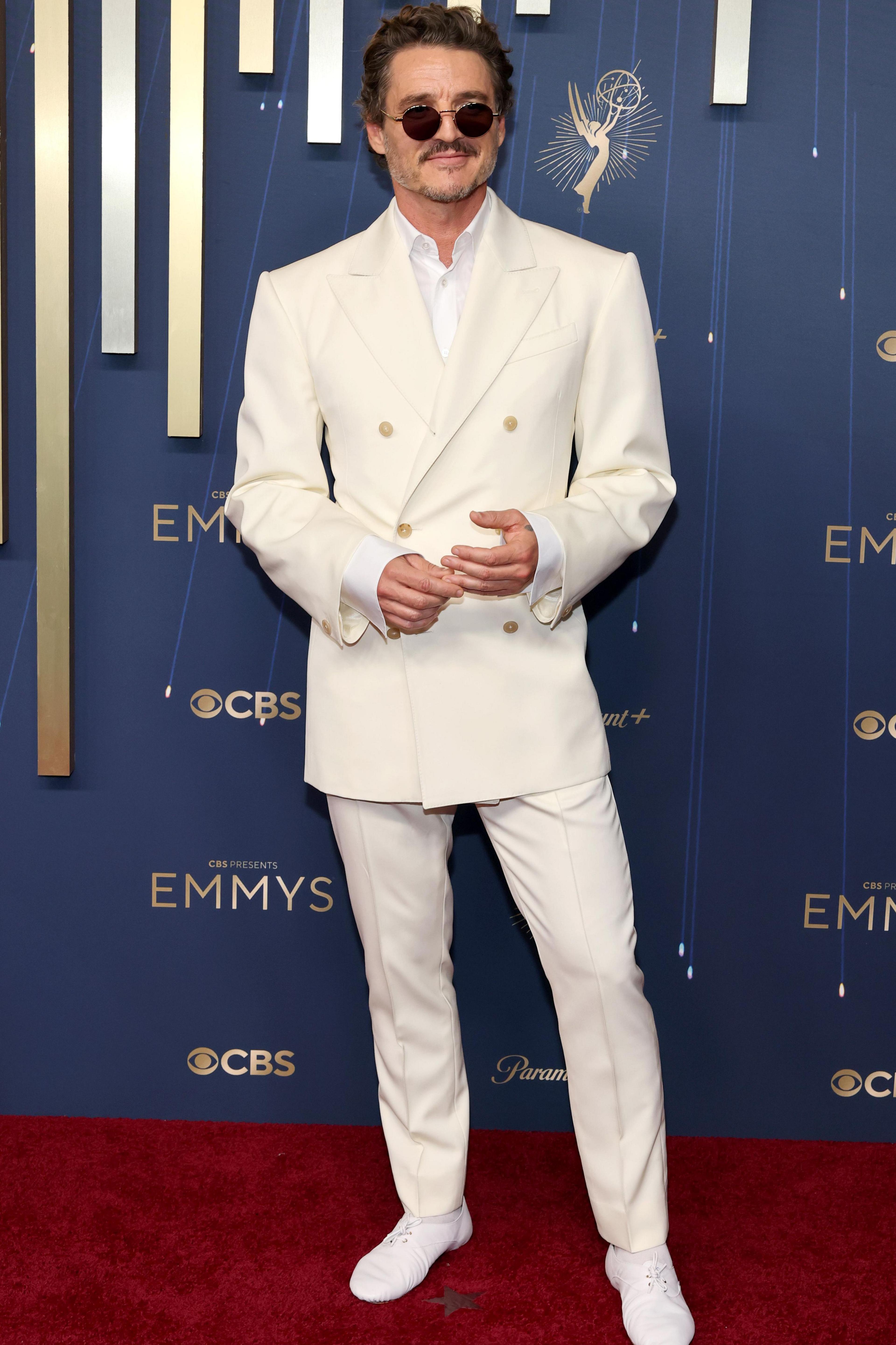 Pedro Pascal looks through brown aviator sunglasses as he poses for photos in an all white suit and shirt. He attends the 77th Primetime Emmy Awards at Peacock Theater on September 14, 2025 in Los Angeles, California.