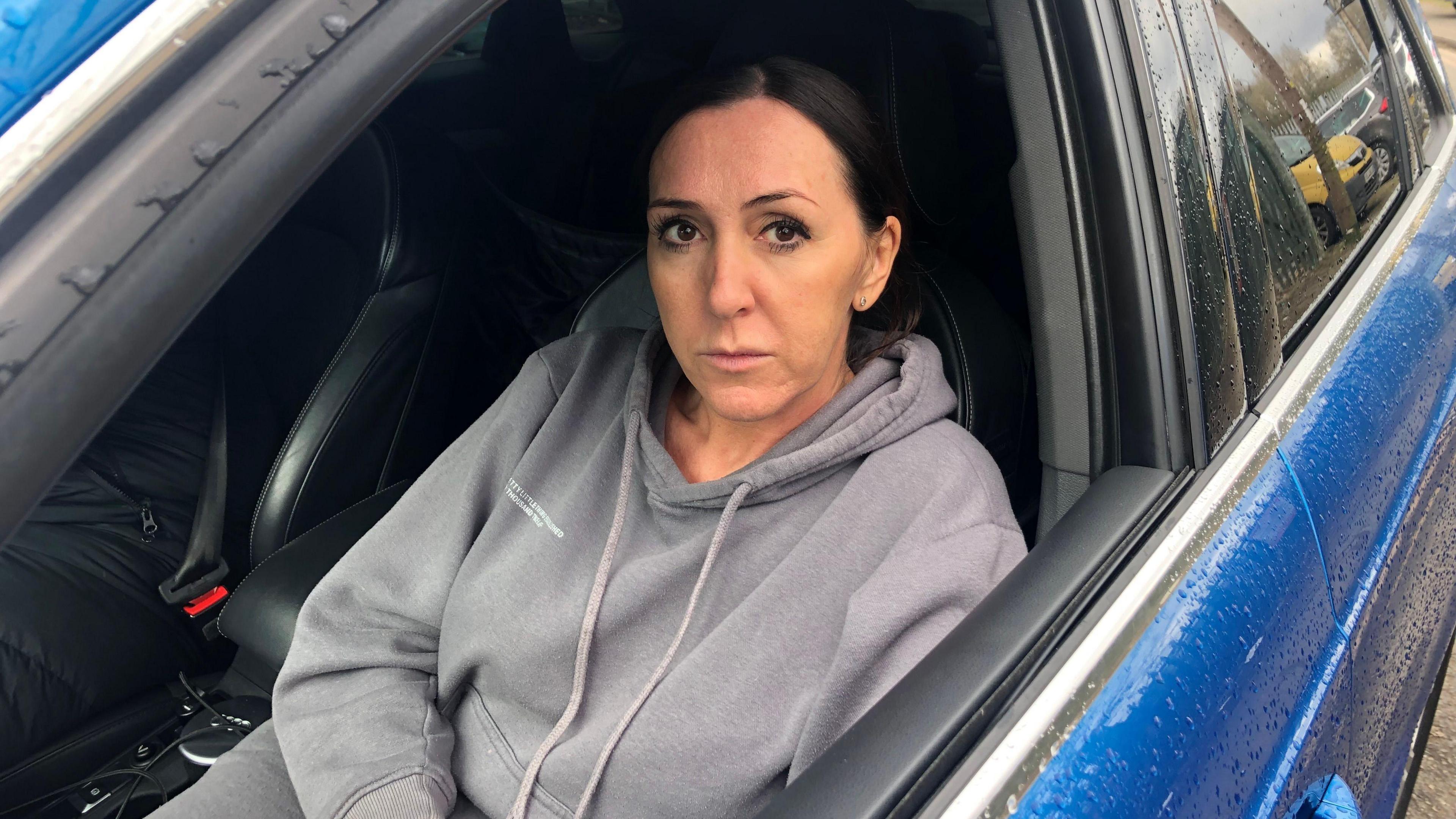 Katrina Devlin in a car. She is wearing a grey hoody. The car is blue. She is looking into the camera. She has blue eyes and is wearing neutral make-up. 