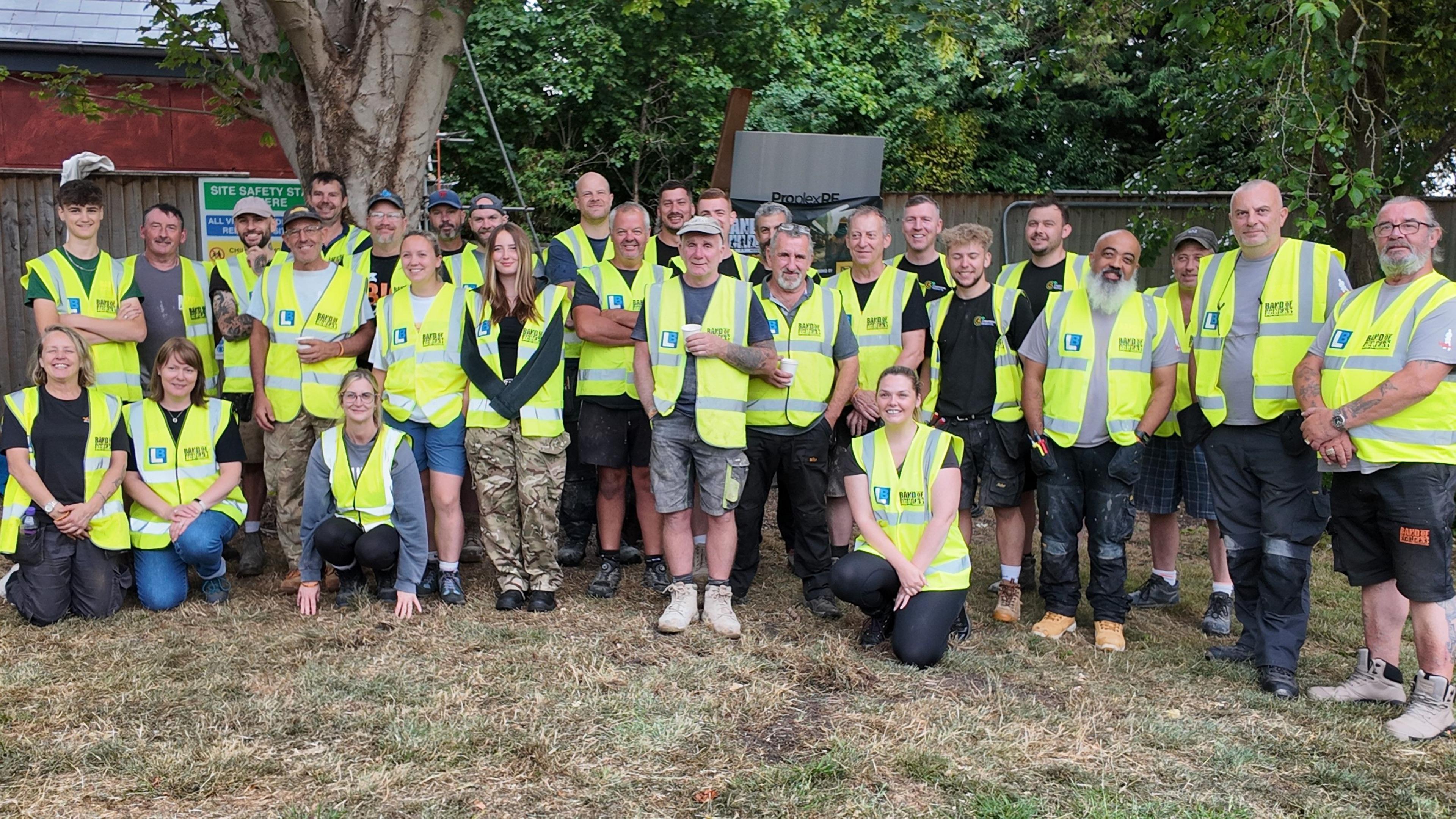 A group of 30 construction workers, both men and women, wearing hi-vis jackets face the camera in a garden.