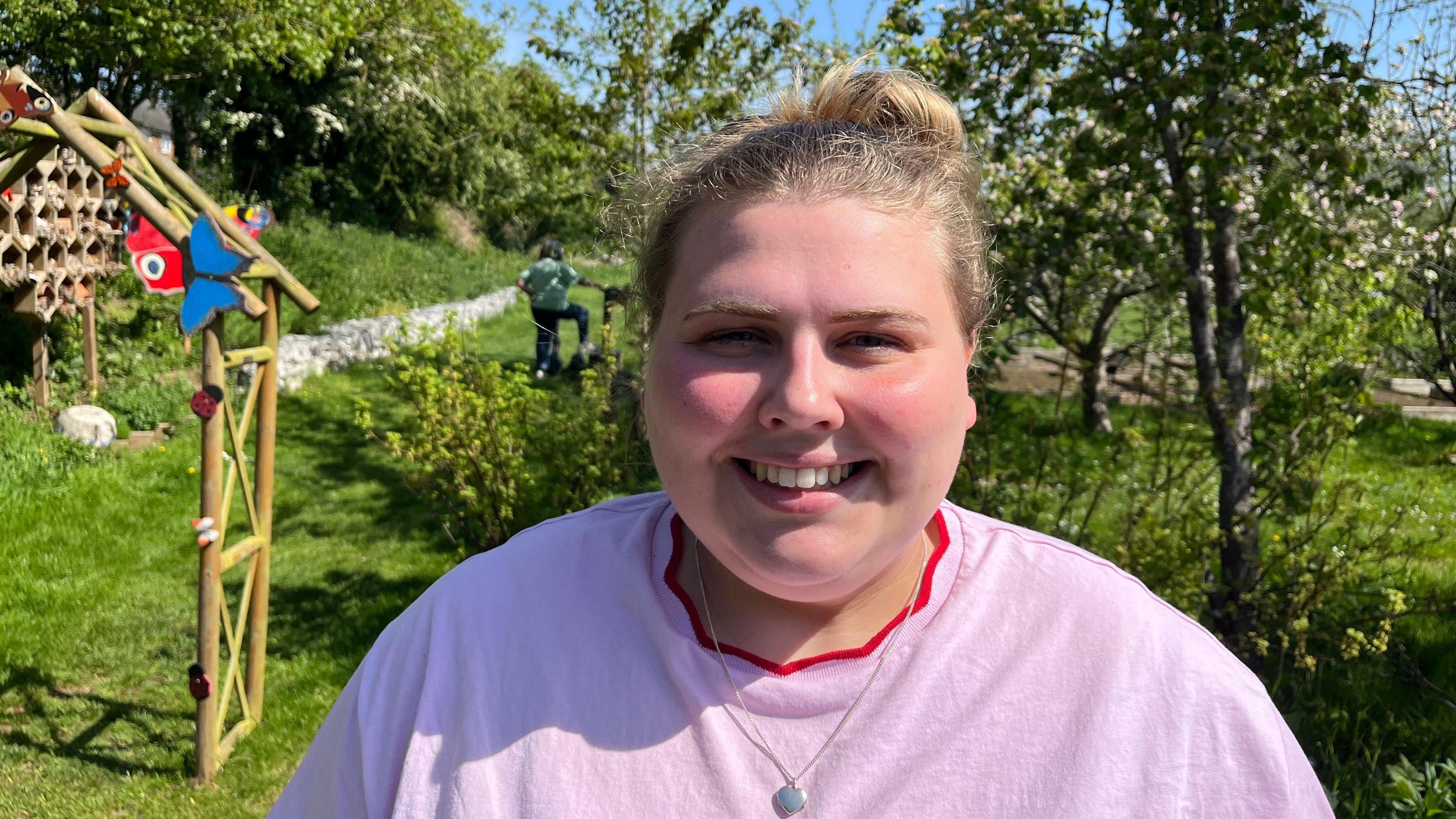 A woman in a pink T-shirt and silver necklace stands in a garden with a wooden structure behind her.