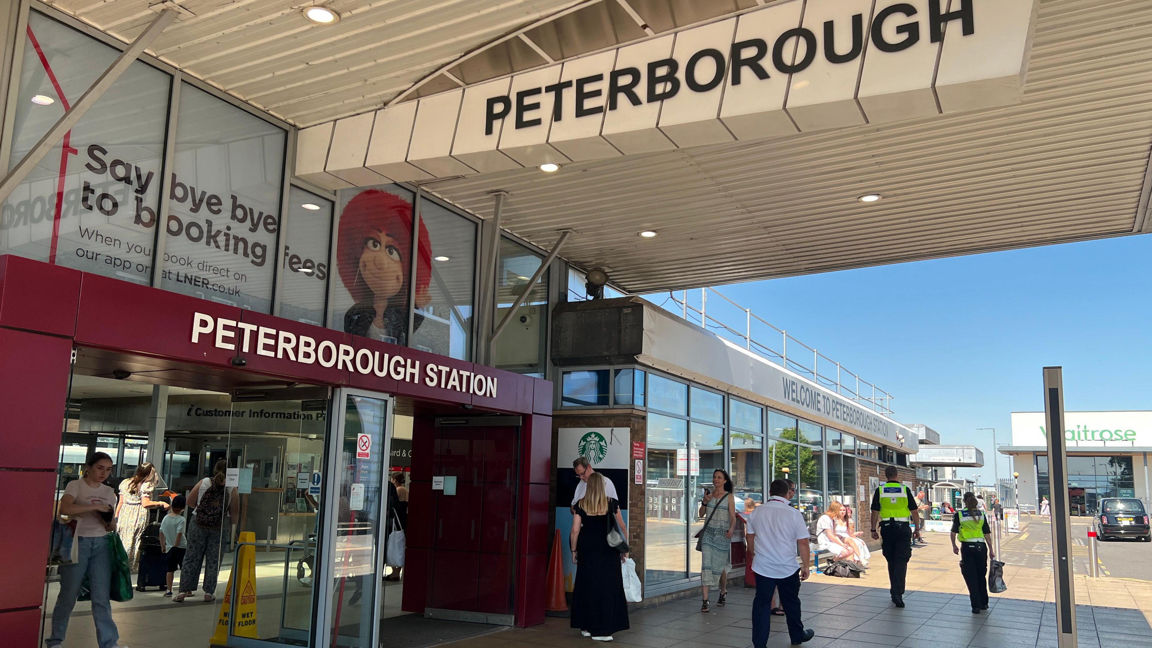 The entrance to Peterborough Station with a large sign above the entrance saying Peterborough Station and men and women walking along the concourse and exciting the station