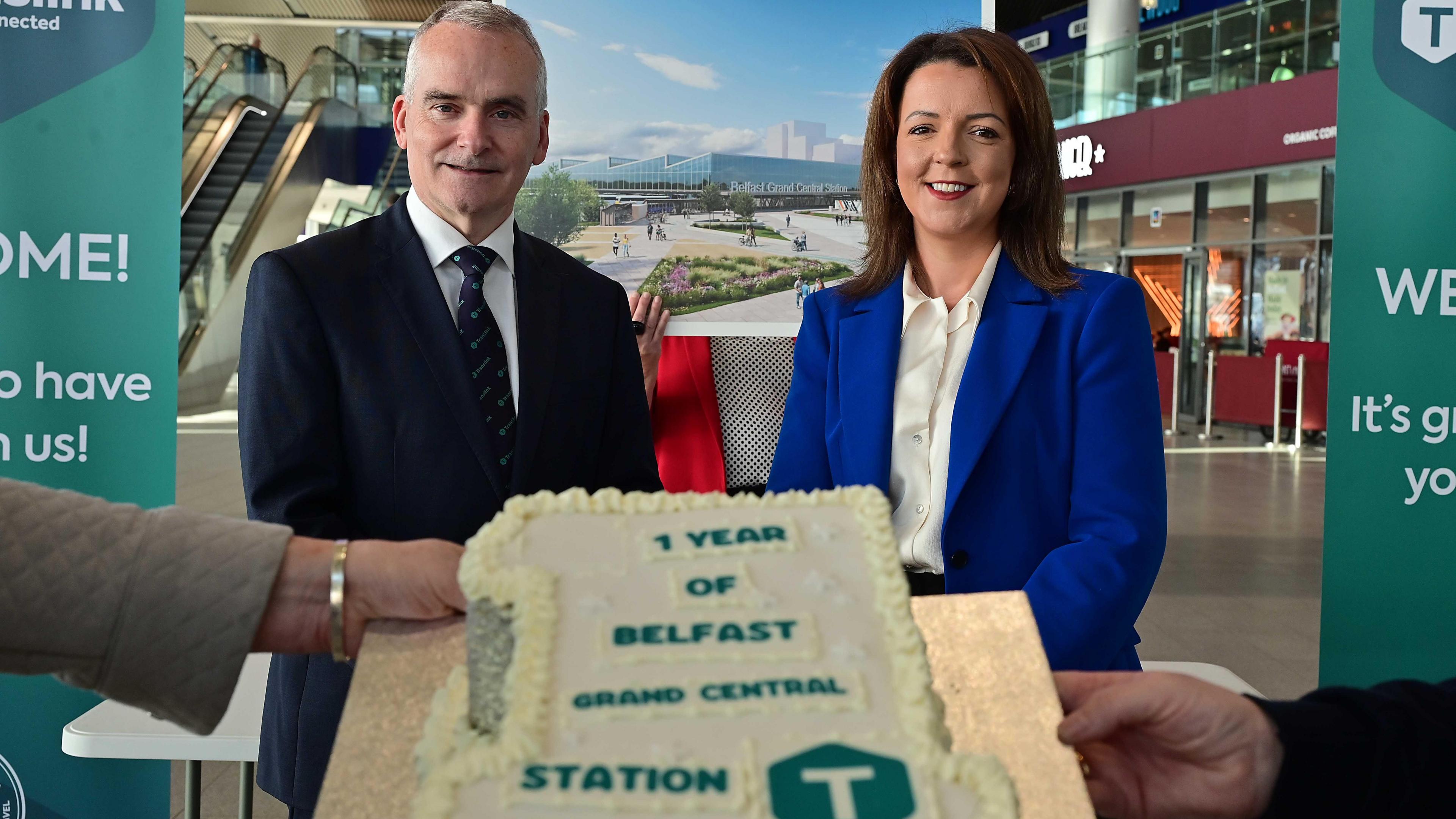 Chris Conway is wearing a suit, Ms Kimmins is wearing a white shirt and blue jacket. Both are smiling at the camera. There is an artist's impression of Grand Central behind them and two hands are seen at the forefront holding up a cake in the shape of a 1. It says 1 year of Belfast Grand Central Station.