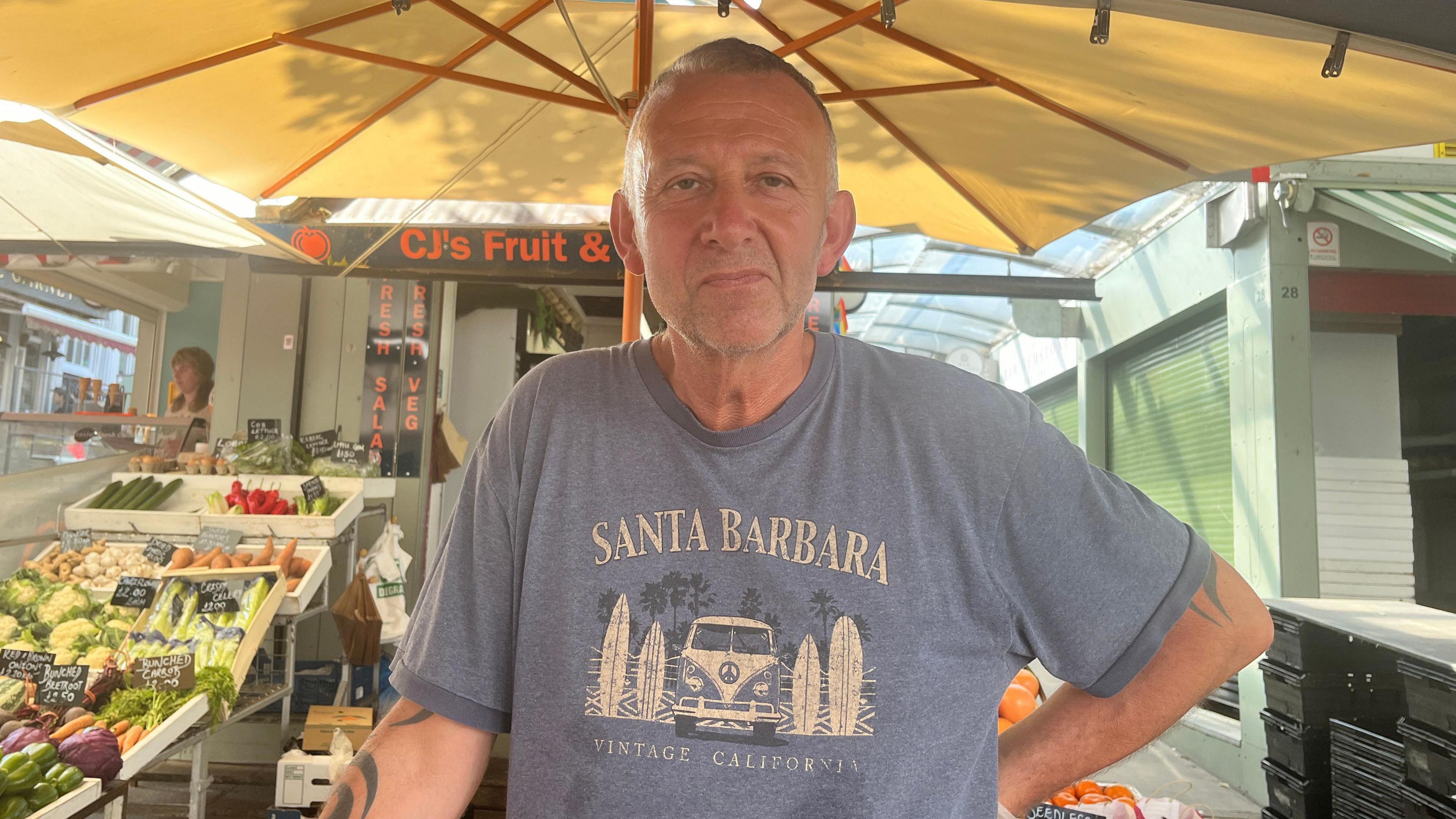 Paul Wiley looks directly at the camera wearing a blue T-shirt, a with a fruit and vegetable stand behind him