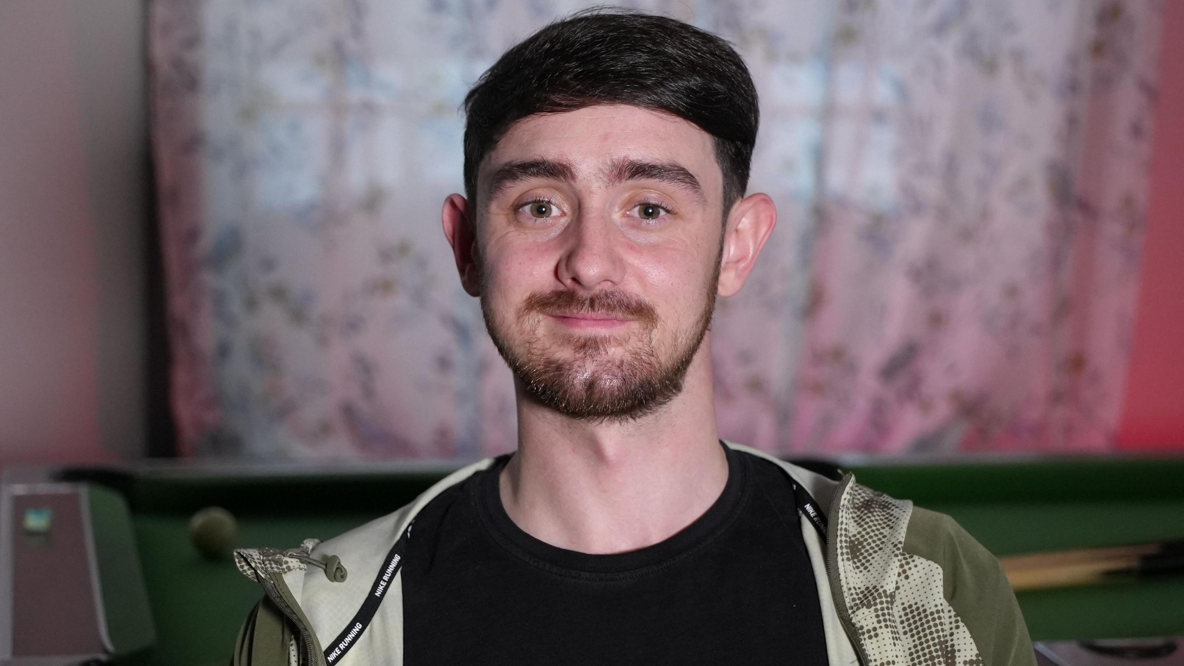 Llun pen ac ysgwyddau o ddyn yn ei 20au yn edrych at y camera ac yn gwenu ychydig. Mae ganddo wallt brown tywyll byr, aeliau tywyll amlwg a llygaid brown. Mae ganddo farf a mwstas brown byr ac mae'n gwisgo crys-t du gyda siaced tracsiwt batrymog wyrdd ar agor dros y crys-t. Mae bwrdd biliards i'w weld y tu ôl iddo, ac mae'r llenni gwyn patrymog wedi'u tynnu ar draws y ffenestr y tu ôl i hynny.