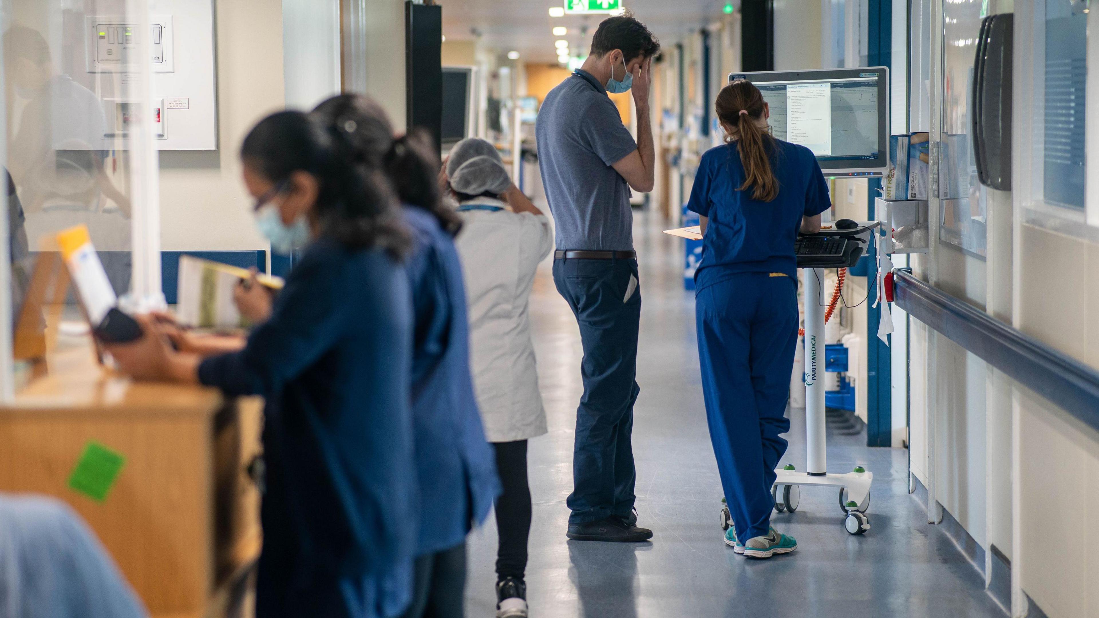 A hospital corridor with staff facing away from the camera. There are two female nurses working at a work station, a tall man in Doctor's clothes rubbing his head as though tired, and another nurse (female) talking to him while accessing data from a computer.