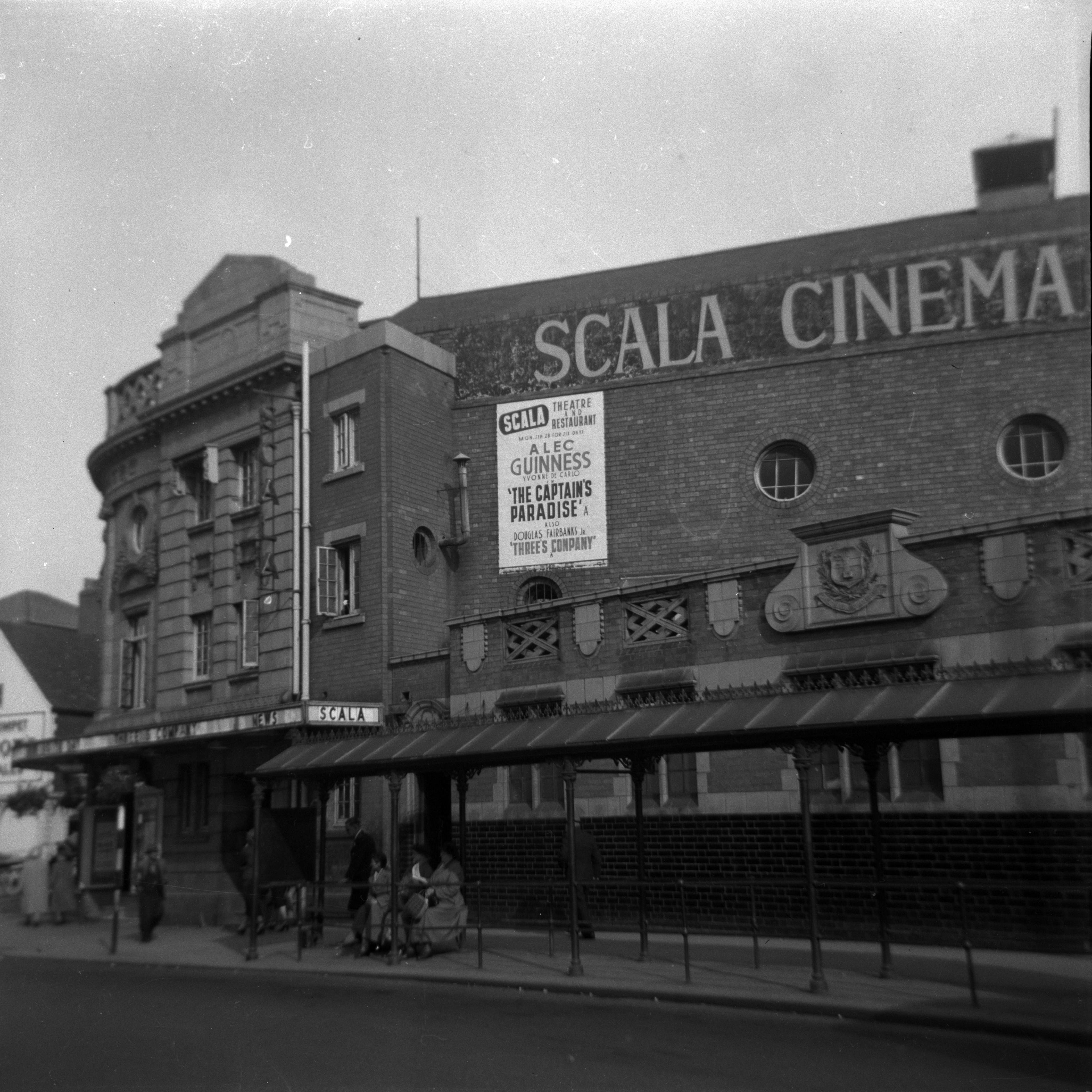 A black and white photograph of the Scala in Worcester as it used to be. "Scala Cinema" is printed in large letters high up on the side of the building. A sign is informing people that The Captain's Paradise, starring Alec Guinness, is currently showing.