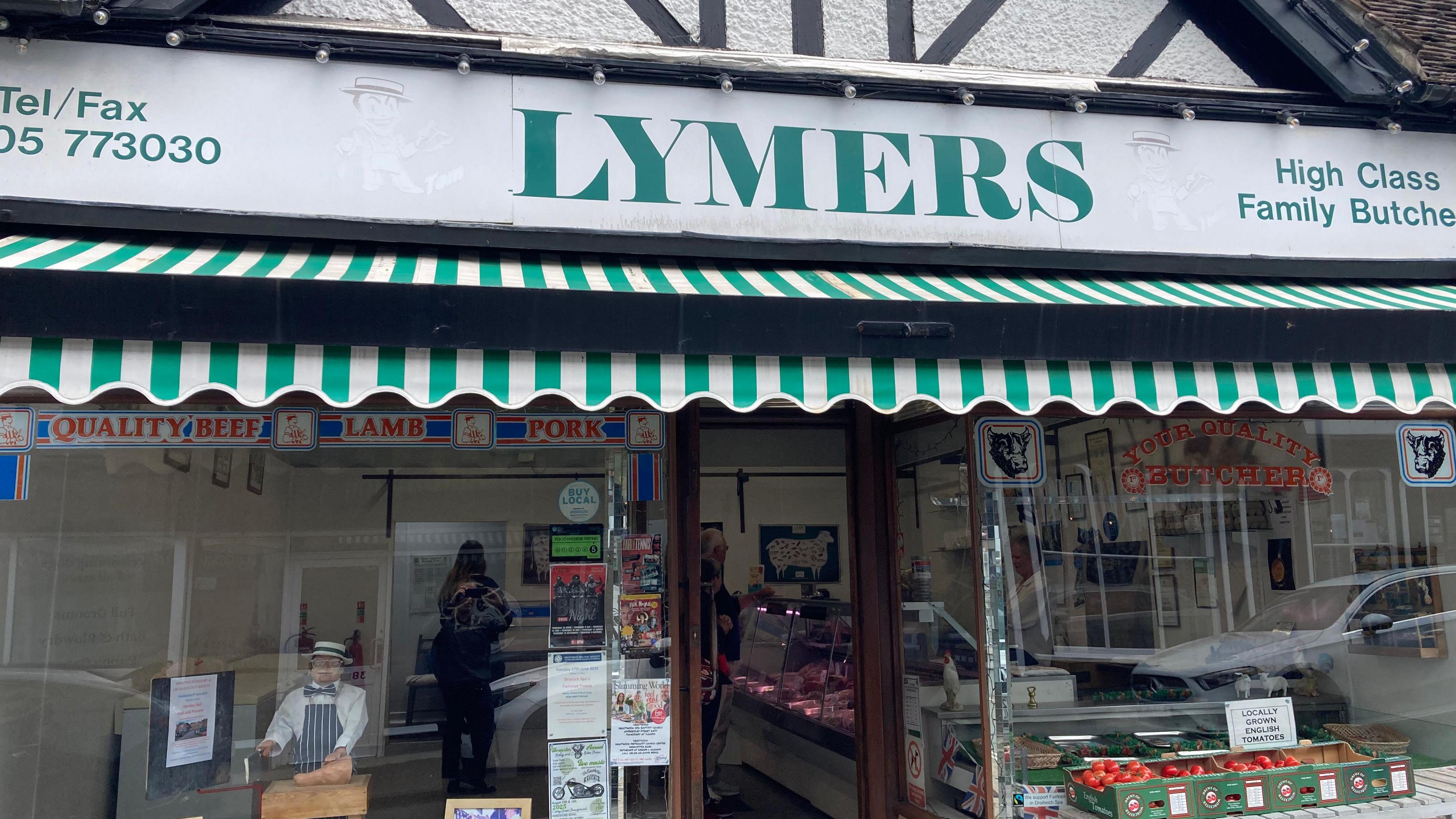 A shop front with a large sign across the top, coloured white and green, with the word 'Lymers' on it - there is also a box of locally grown tomatoes on display.