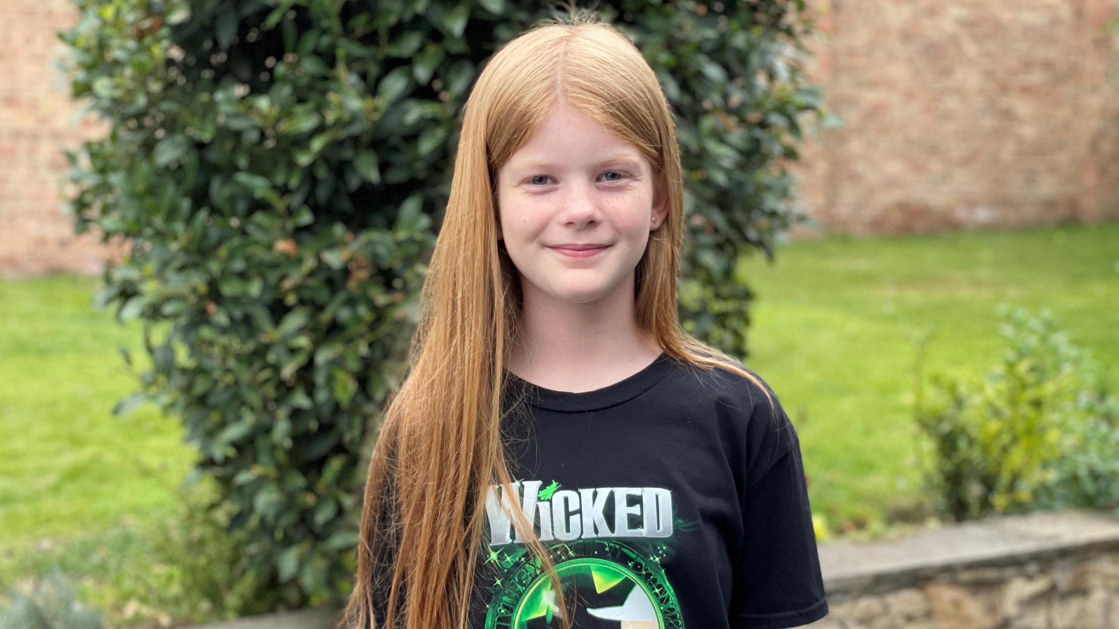 A girl with red hair stands in front of a green bush in a garden. She is wearing a black t-shirt with the Wicked stage show logo. There is a garden wall in the background. 