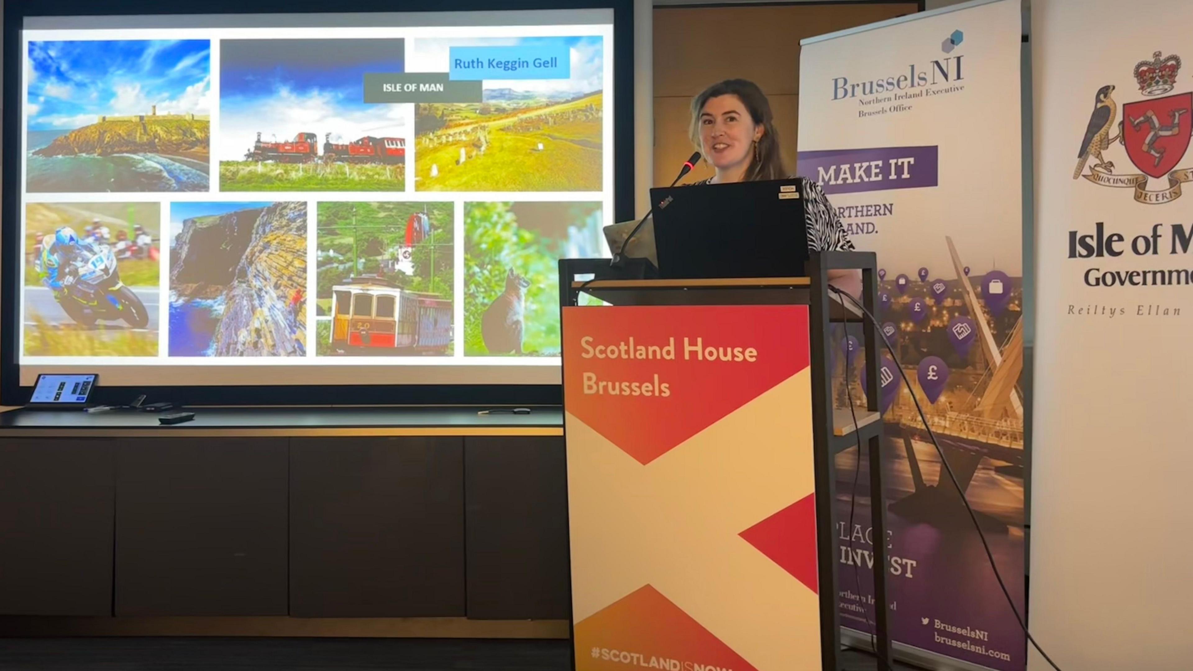 Ruth Keggin Gell standing at a lectern with a power point presentation on screen next to her that includes images of several Manx attractions, including the Manx Electric Railway, Isle of Steam Railway, TT motorcycle racer, a wallaby and Peel Castle.