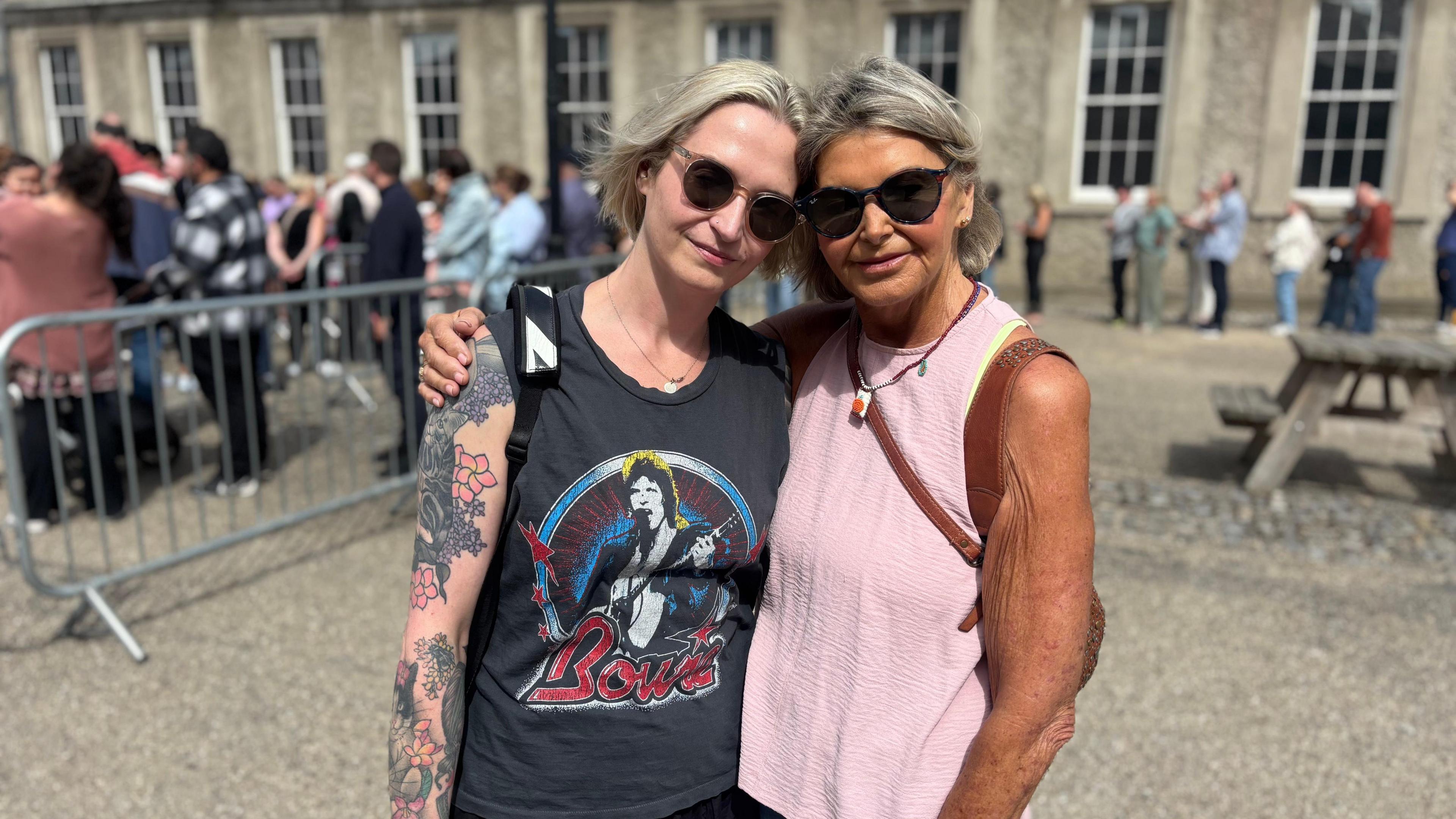 Two women pose for a photo, both are wearing best tops, on the left the woman has light hair and a navy top with a David Bowie design, on the right the woman has grey hair and is wearing a pink top.