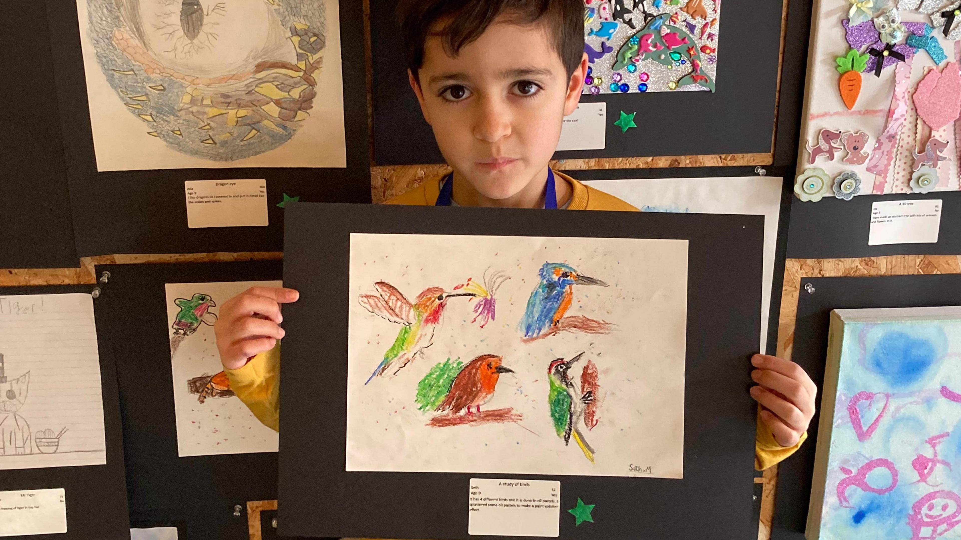 A young boy holding up a picture of four different birds, standing in front of a wall of paintings.