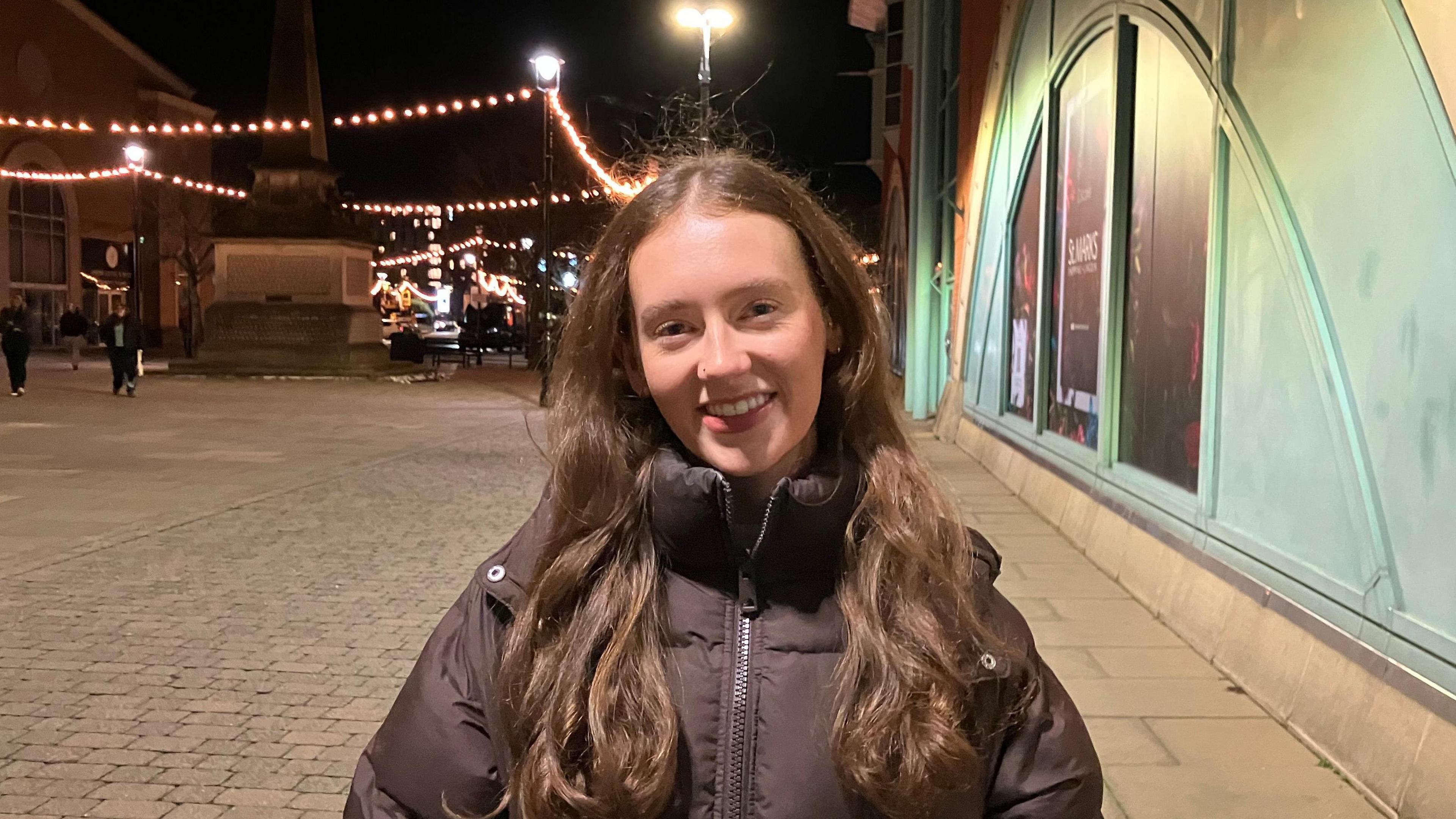 A head and shoulders shot of Annie-Mae Haines smiling at the camera. She is standing outside a shopping centre. She has long brown hair and is wearing a brown coat.