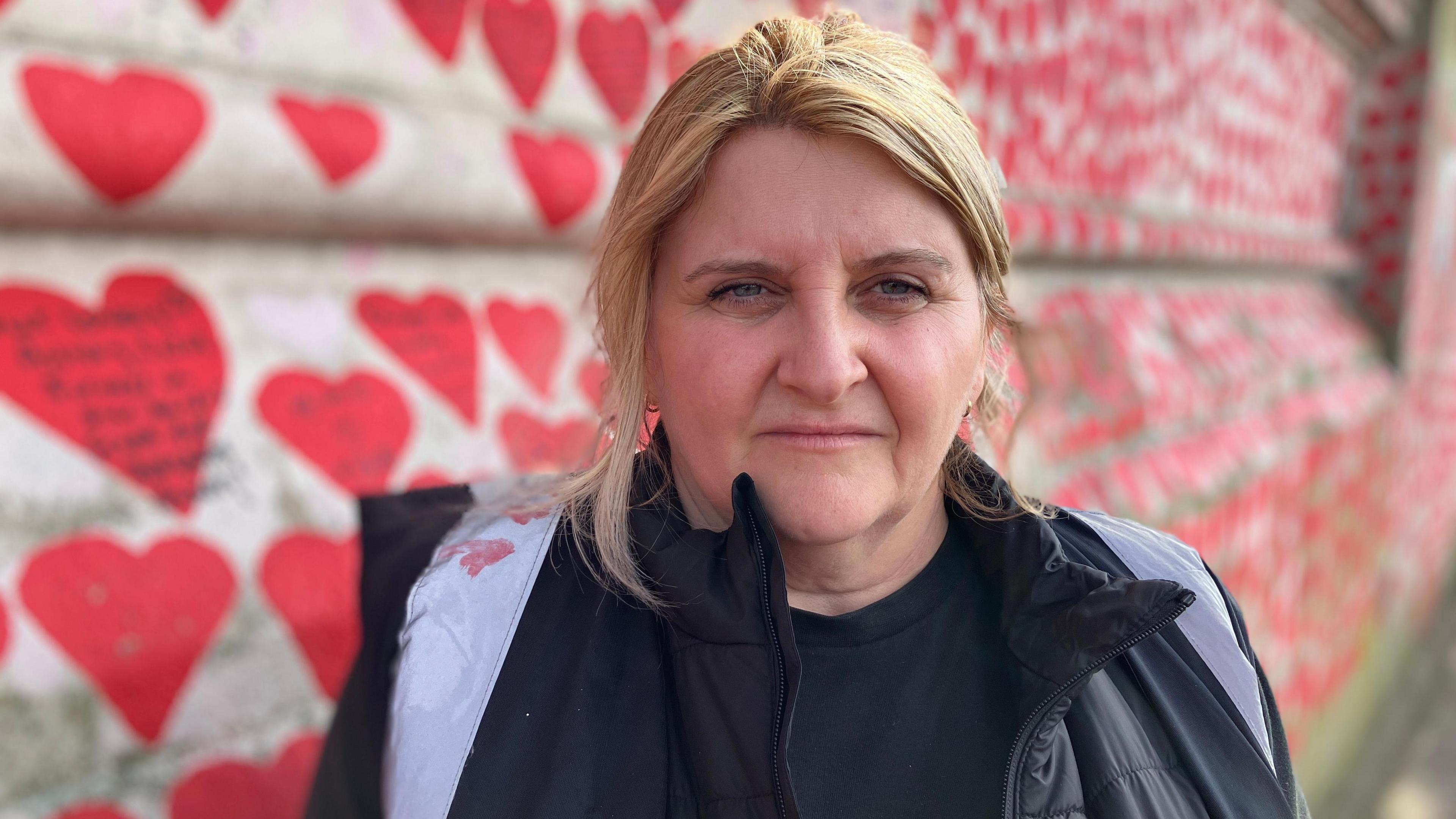 A woman with blonde hair looks into the camera with a serious expression, in front a wall painted with lots of red hearts