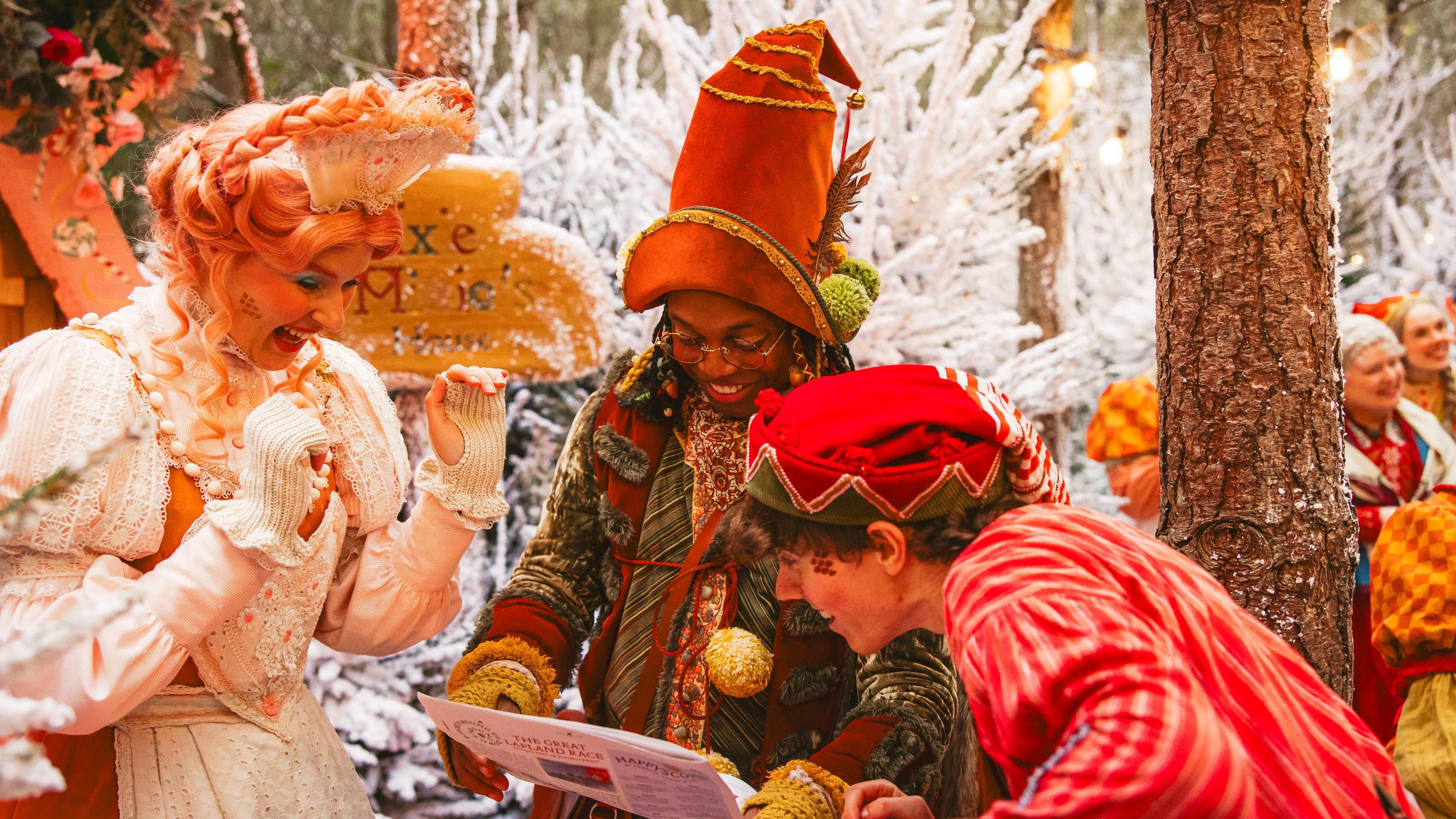 Cast members at Lapland UK - a woman in a white traditional Christmas dress with long plait is looking at a newspaper with a smile on her face. The person in the middle has an elf hat on and is holding the newspaper. Another elf looks on and is smiling too. 