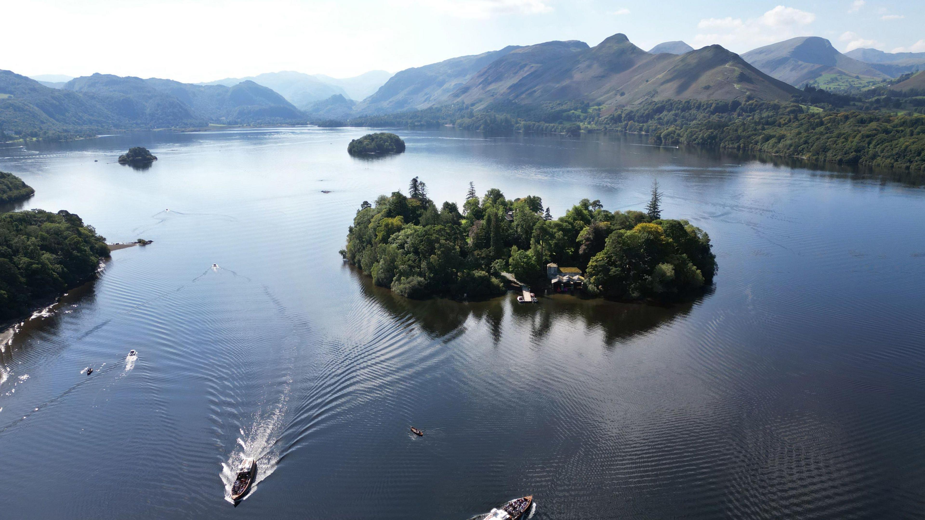 A beautiful sunny day over Derwentwater near Keswick in the Lake District. An aerial view shows islands on the lake and impressive peaks in the distance. In the foreground, there is a scattering of boats of various sizes, some showing a wake behind them.