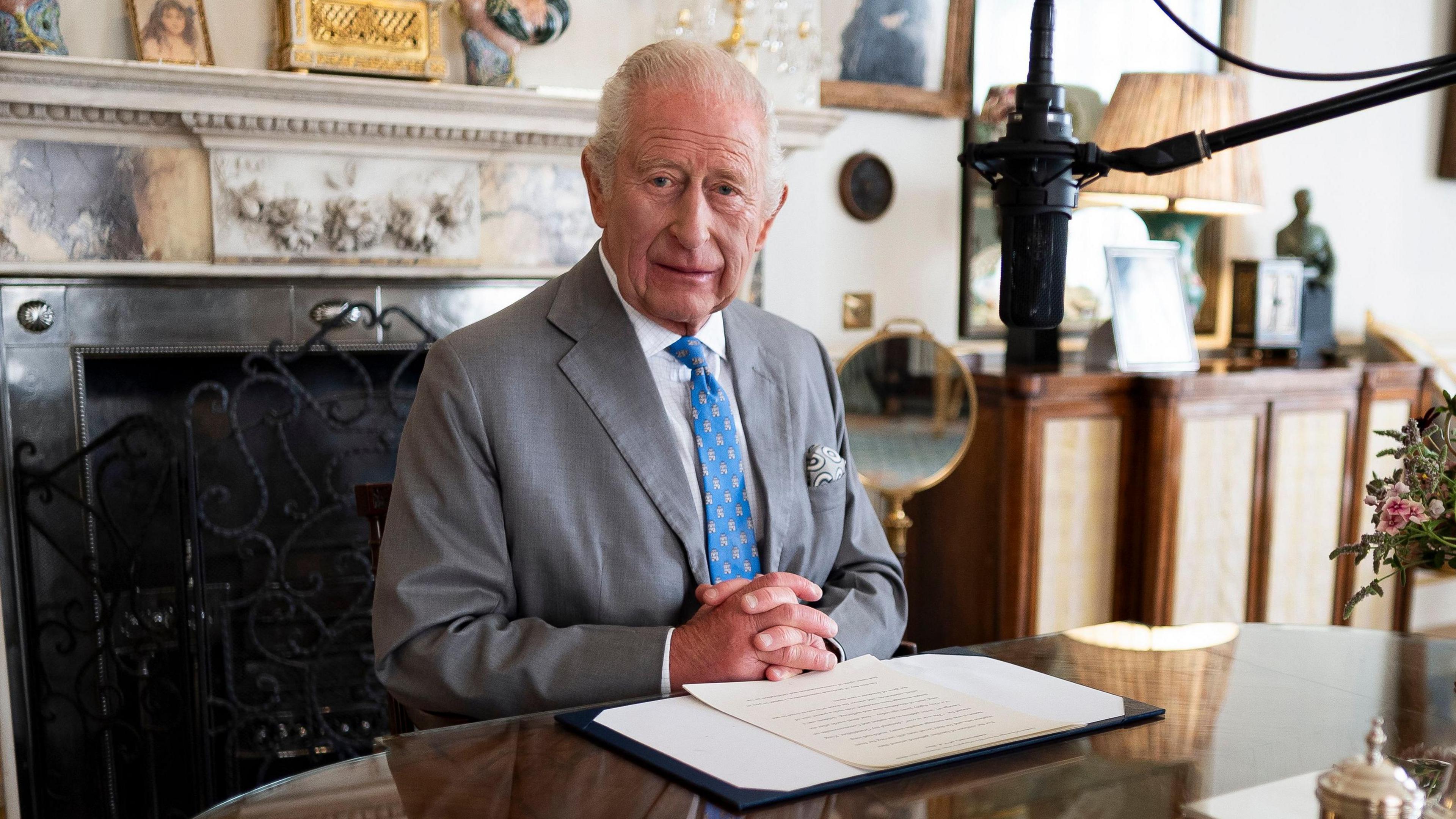King Charles III wears a grey suit with a striking blue tie with his hands clasped at the foot of a piece of paper containing the words of his VJ Day message in the Morning Room of Clarence House, London. He is sitting in front of a marble-style fireplace. A microphone hovers near to this face.