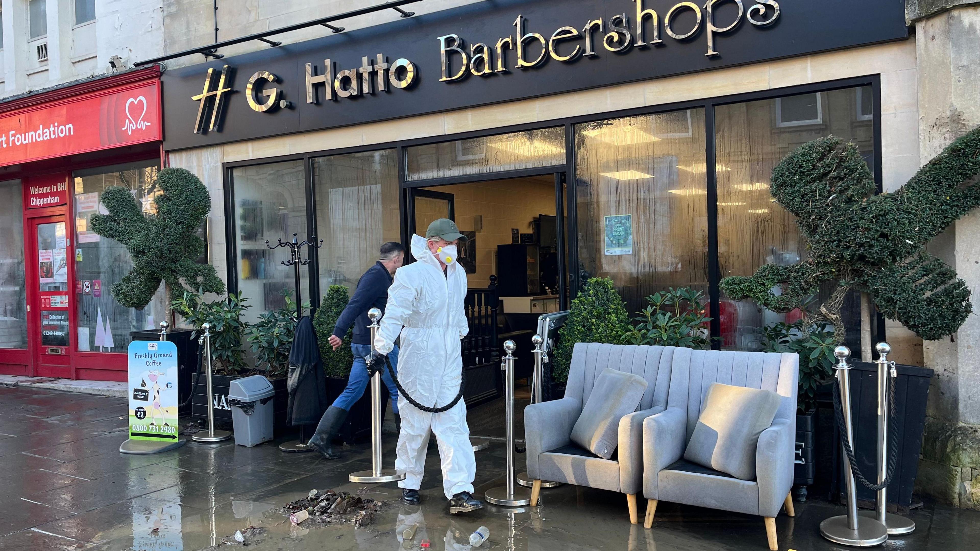 The front of a barber shop which was flooded. Outside are two grey chairs which have been moved out of the shop. There is a man in an all in one white  overall with a mask over his face. Another man, wearing black wellington boots, is entering the shop. 