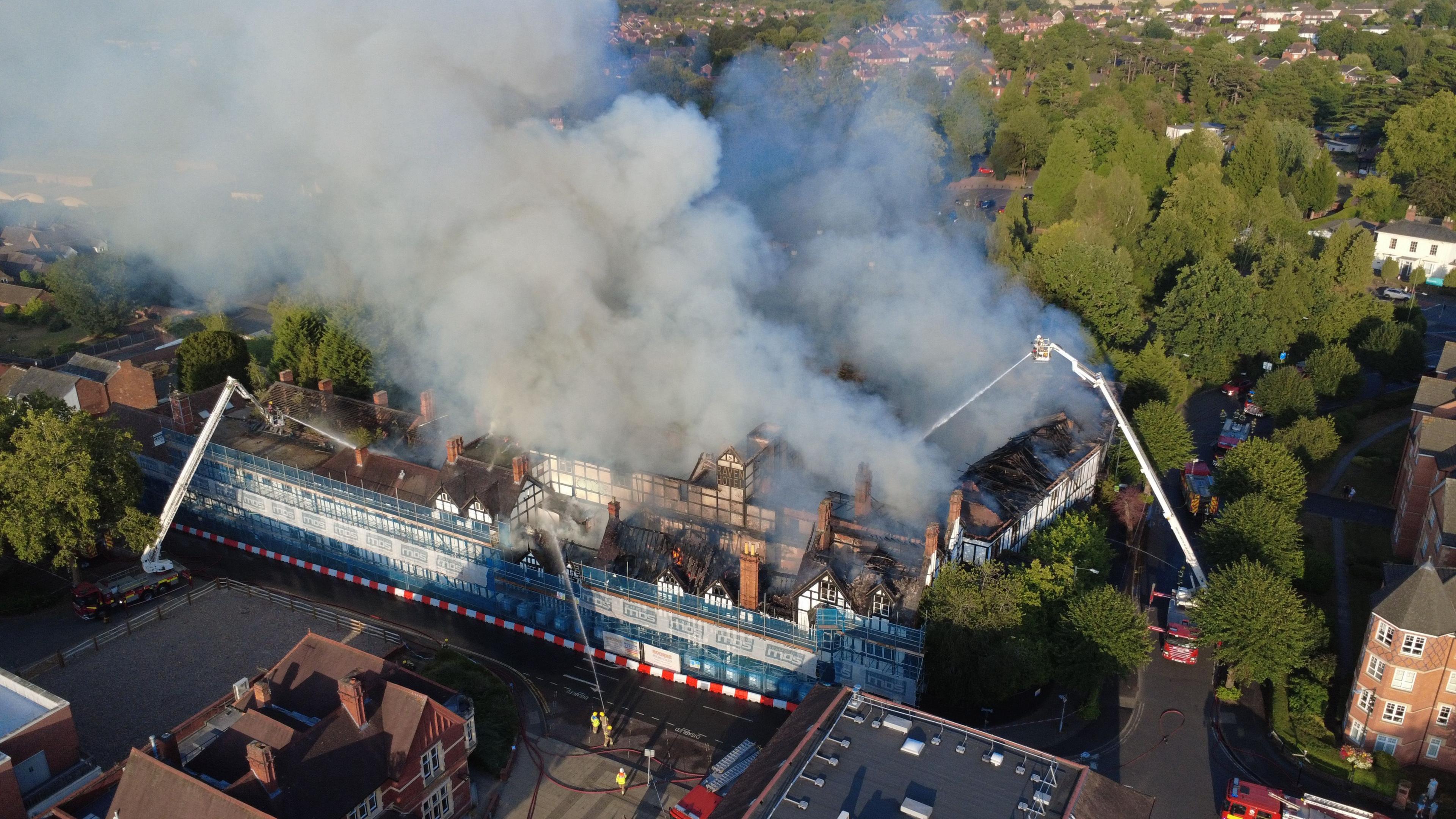 A black and white building engulfed in smoke. There are firefighters on two aerial platforms spraying water on it