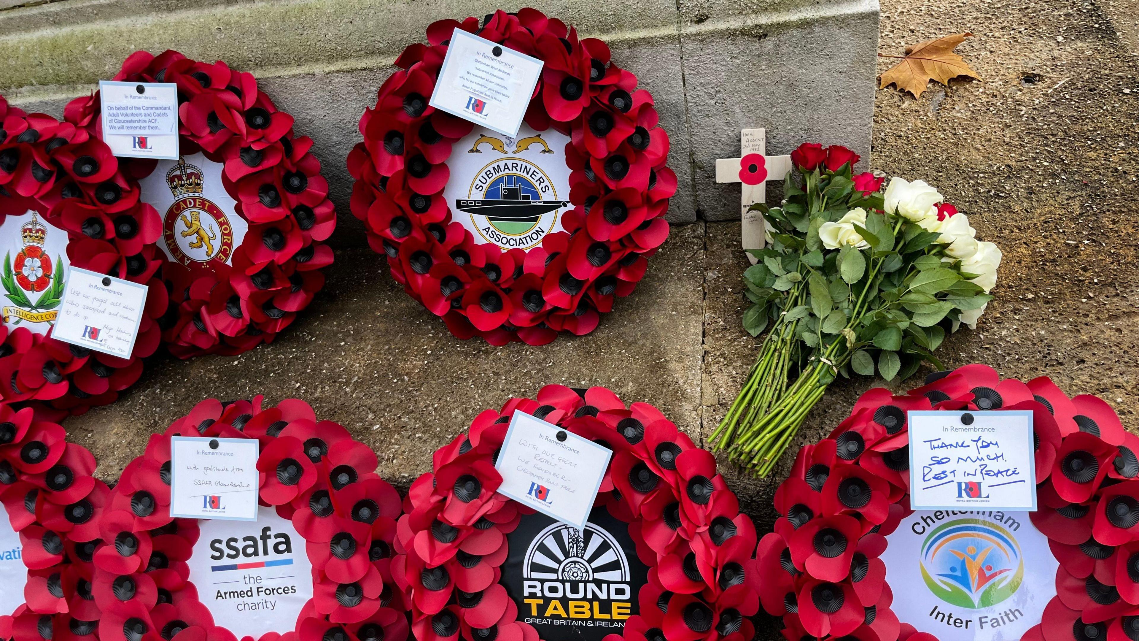 Several poppy wreaths, a bouquet of flowers and a crucifix cross on the ground. The wreaths have notes attached to them and the logo of the organisation they came from.