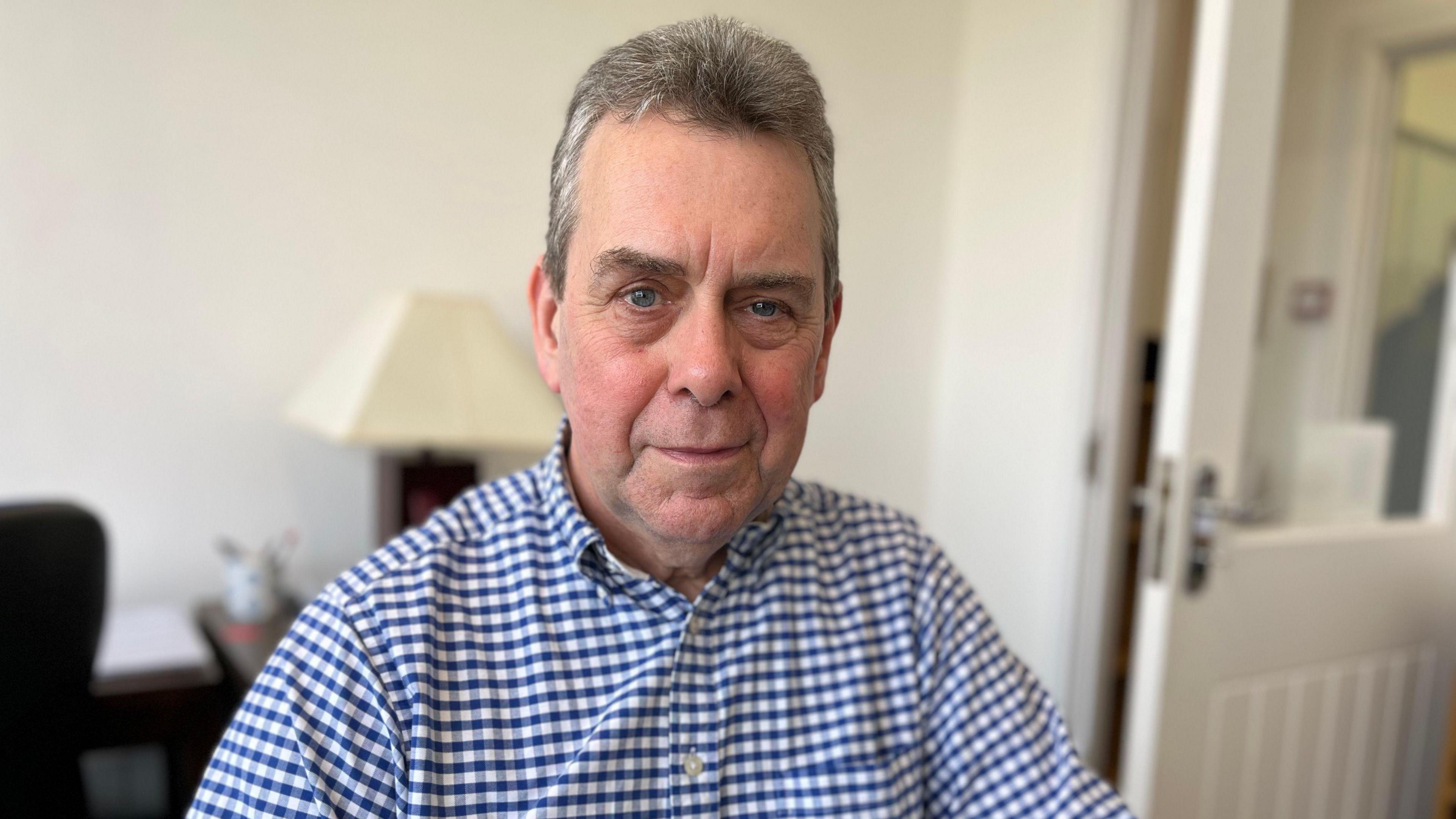 David Thompson has grey hair, blue eyes and is smiling with pursed lips. He's sitting in his office and wearing a blue and white gingham shirt with the top button unbuttoned. In the blurred background is a lamp and a white door. 
