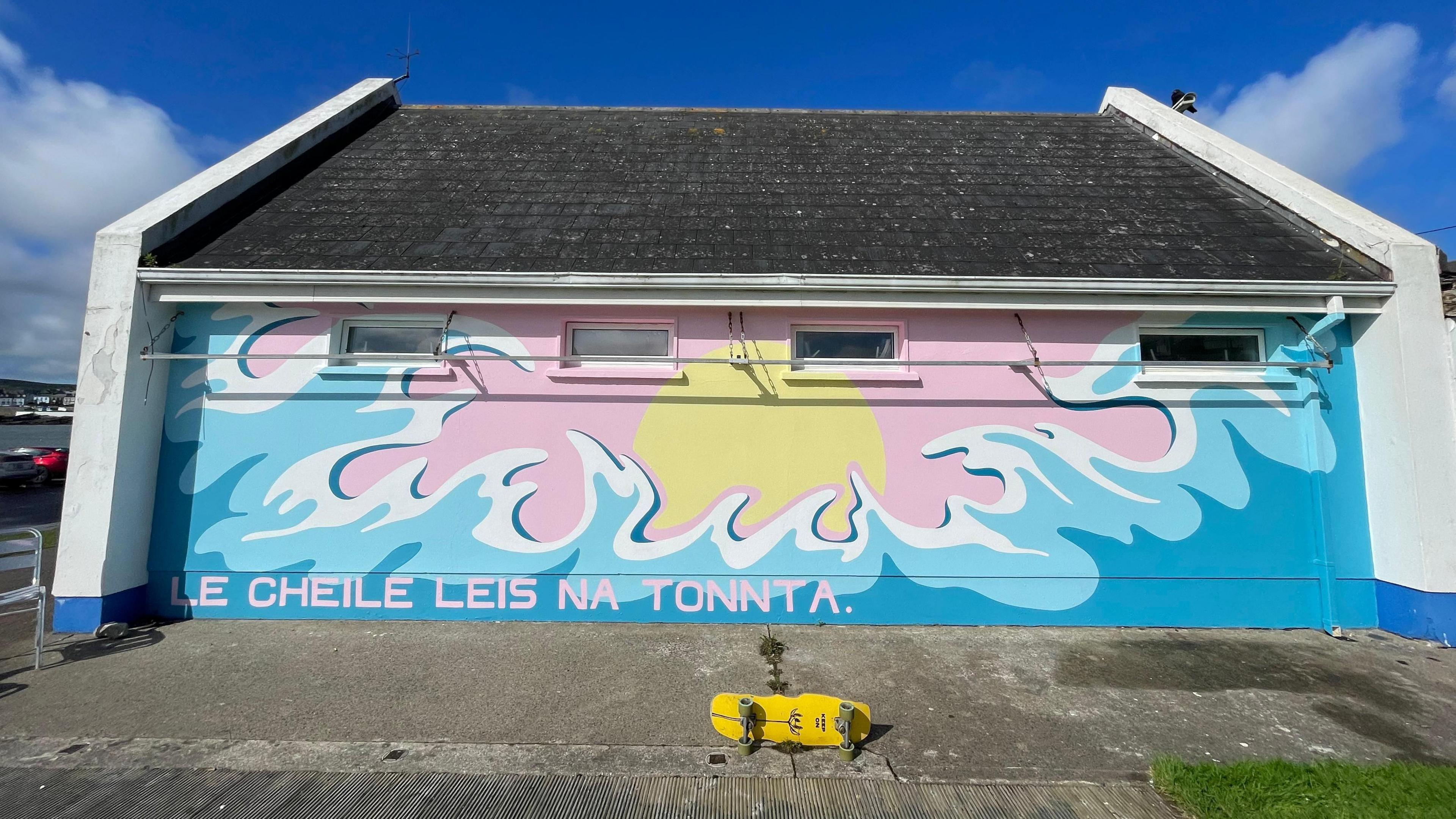 This mural is painted on the side of a one story building. On it, we see a surge of blue waves upwards towards a pale yellow sun, set against a baby-pink sky. In the same colour, there is a message at the bottom left reading le cheile leis na tonnta, which is Irish for together with the waves. On the ground at the bottom, her yellow skateboard is laying down sideways.