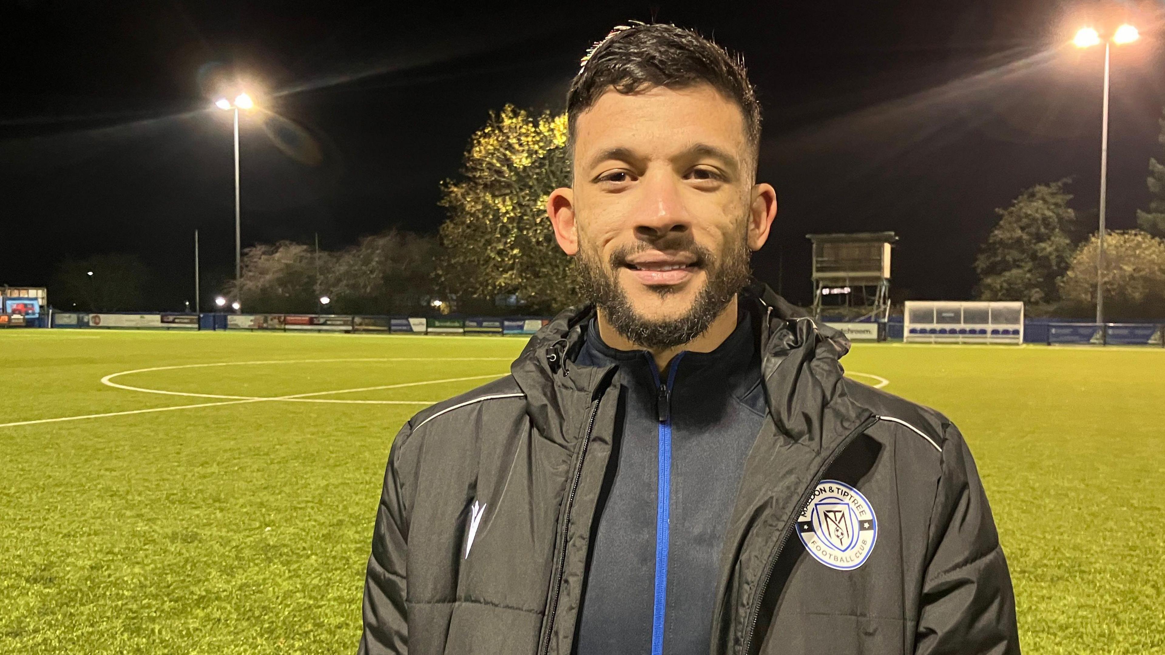 Macauley Bonne at Maldon and Tiptree FC's ground. He wears winter football gear and the pitch is floodlit at night. He has short dark hair and a neat beard and moustache.