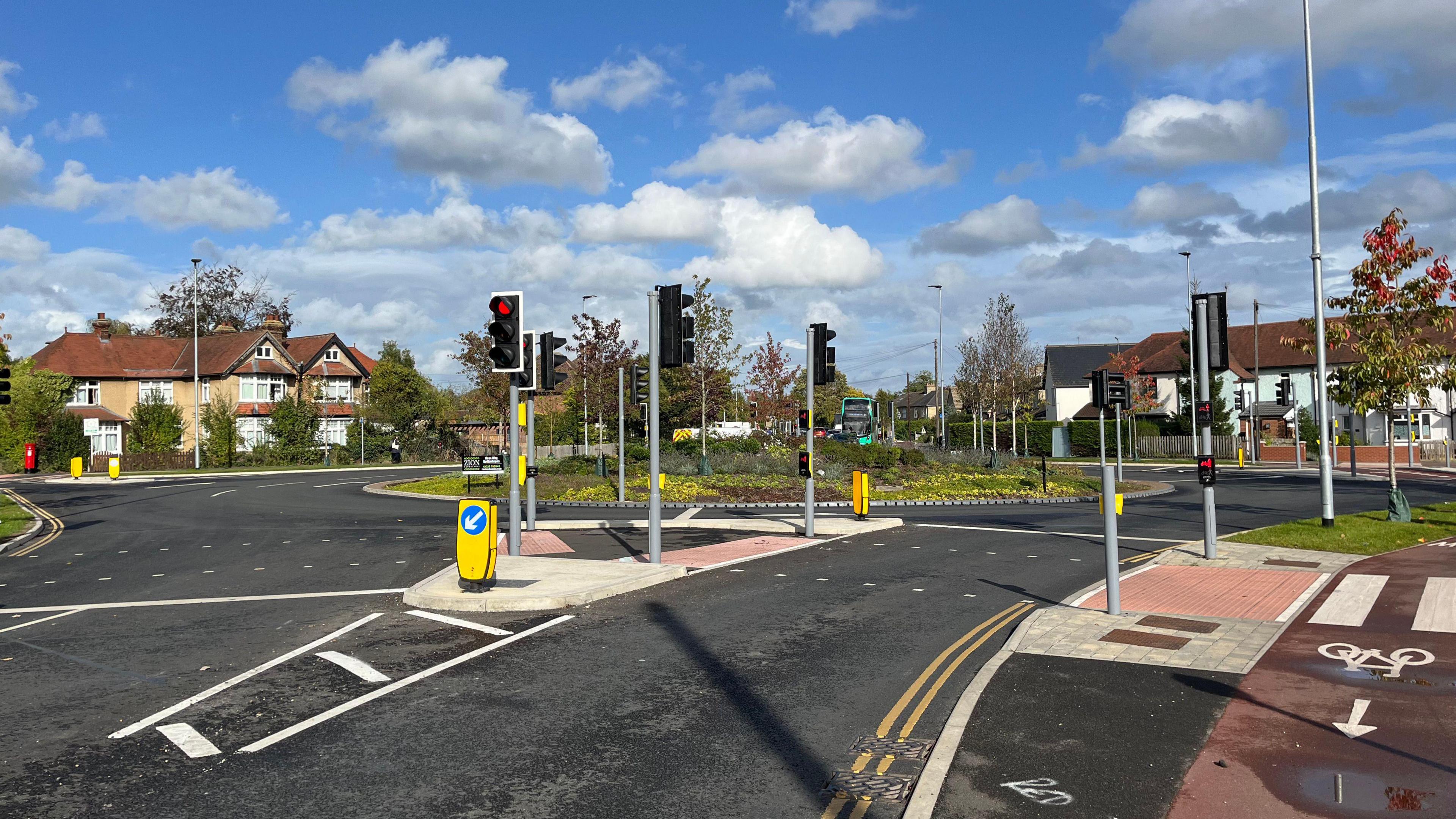 A road approaching a roundabout with a pavement, traffic lights and houses and greenery in the distance.