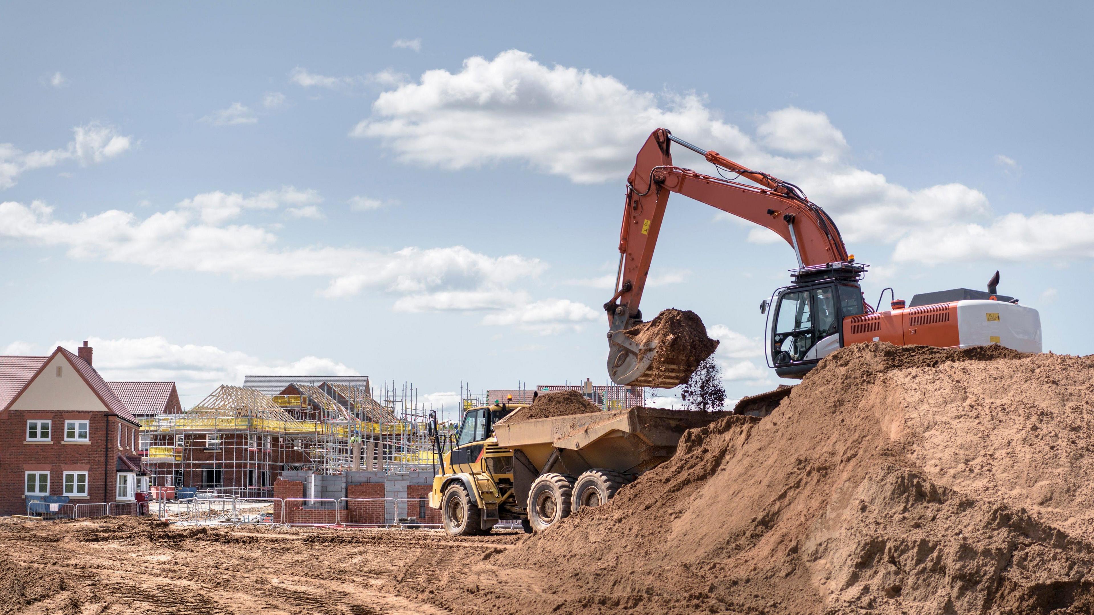 A stock image of a construction site. A digger can be seen on top of a large pile of earth, lifting it into the back of a truck. In the background there are new-build houses, some of them covered in scaffolding. 