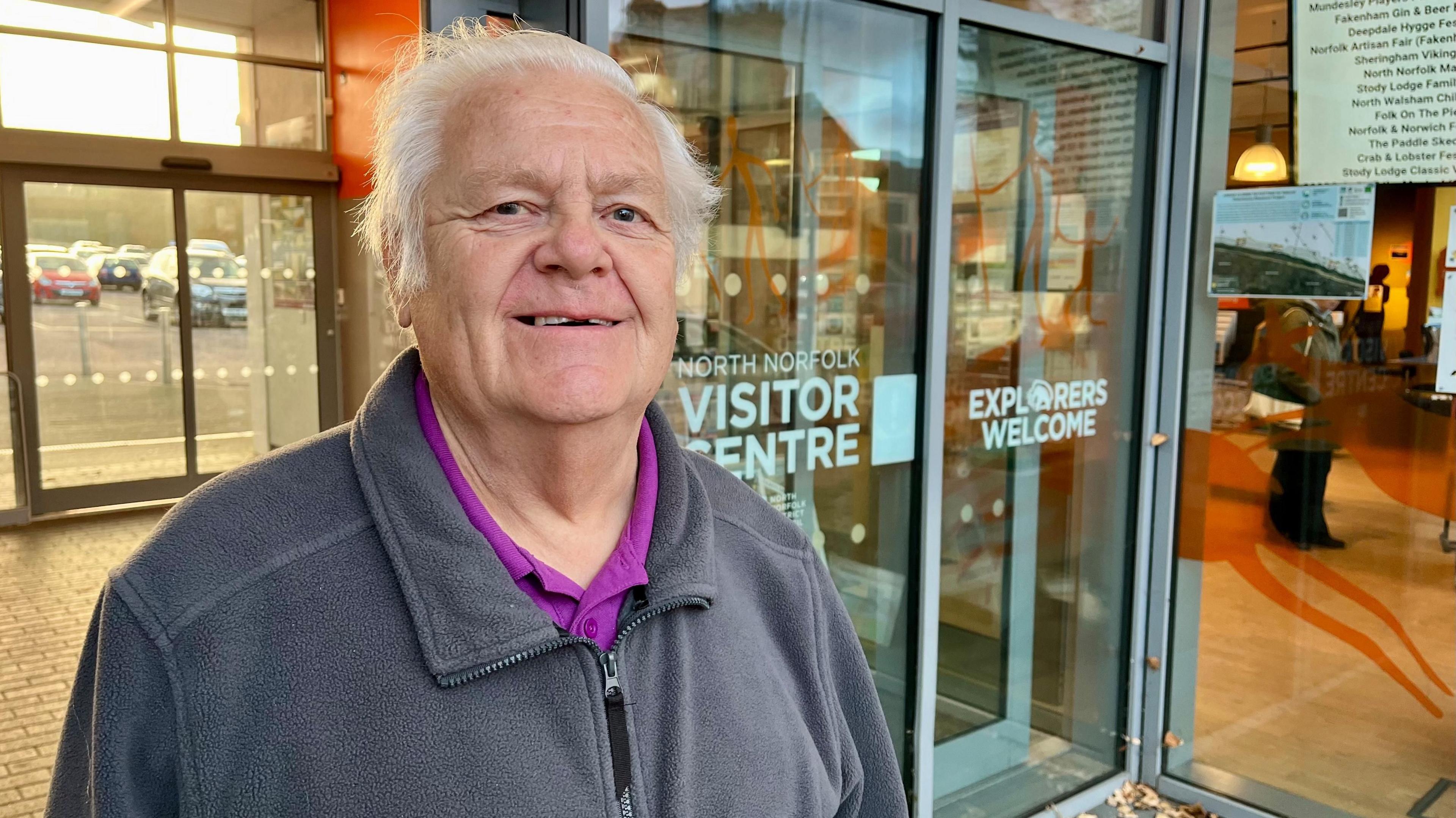 Derek Simon is standing in front of the door to the visitor centre.
He has short, white hair and is wearing a grey fleece jacket with a purple top underneath.
The visitor centre shop can be seen through the window behind him.