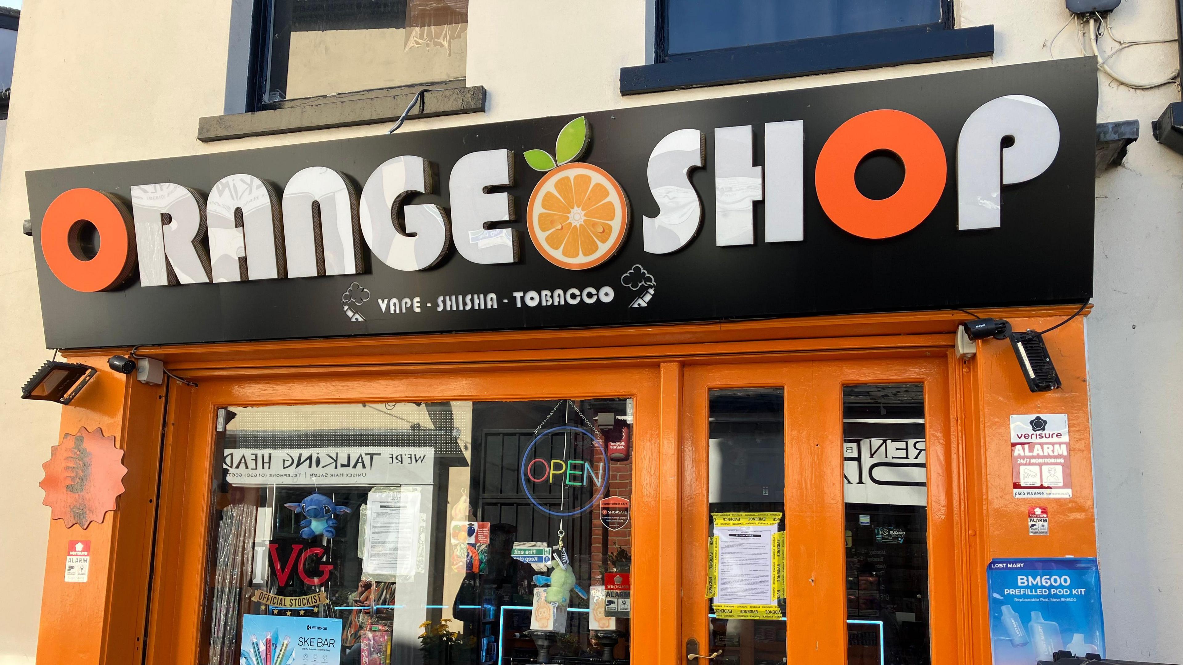 A shop with a sign that reads Orange Shop, Vape, Shisha, Tobacco. There are posters and items in the window. 