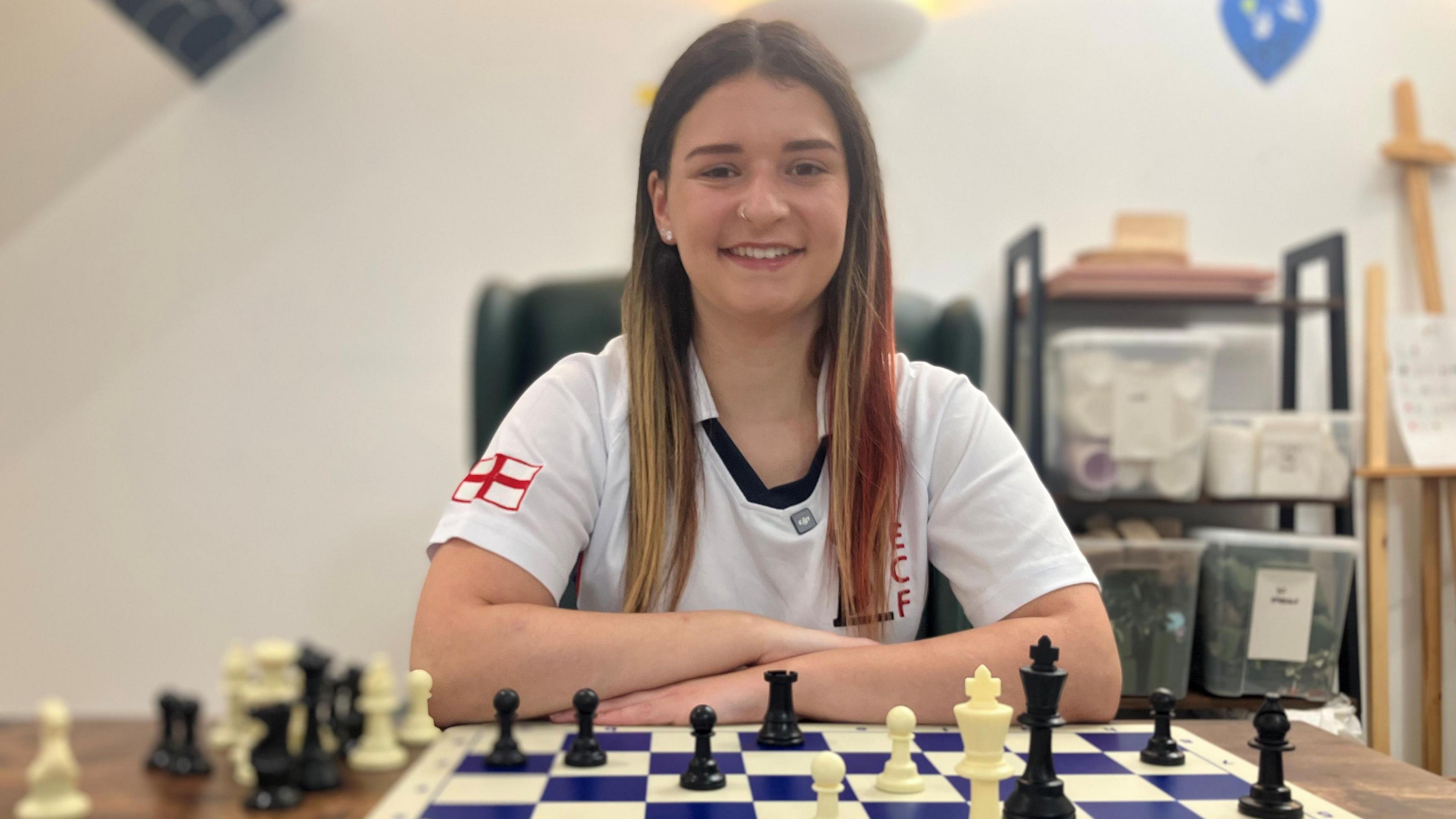 A white teenage girl with long light-brown hair with red streaks sitting at a table with a blue and white chess board and white and black chess pieces on it
