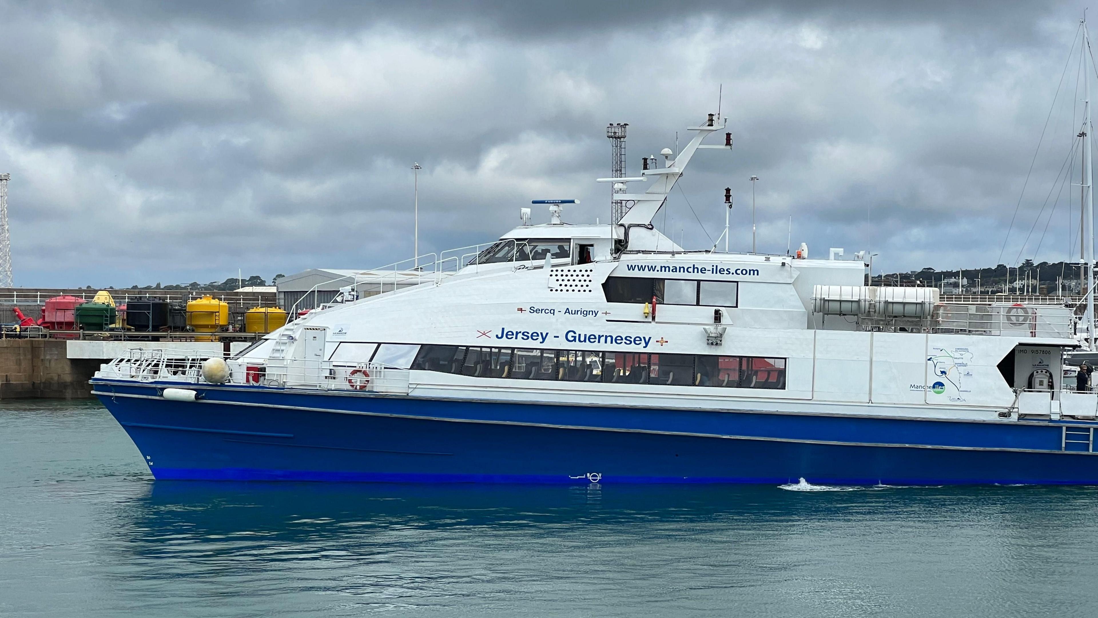 A blue and white boat branded www.mache-iles.com and with Jersey-Guernsey written on its side is seen in a body of water with a pier behind. 