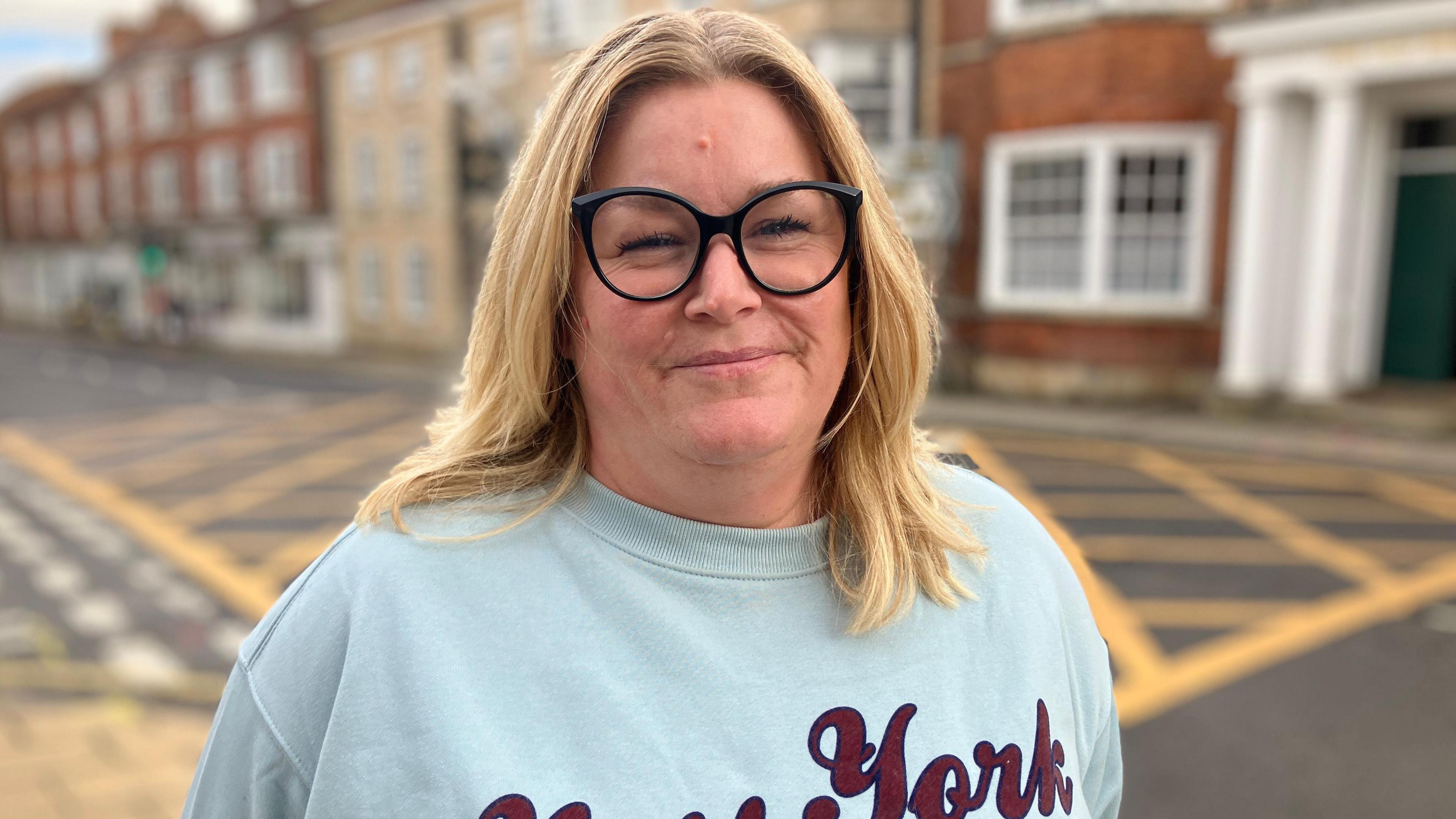 A woman with blonde hair and thick black-rimmed spectacles pictured on a town street. A yellow box junction and terraced properties can be seen behind her.