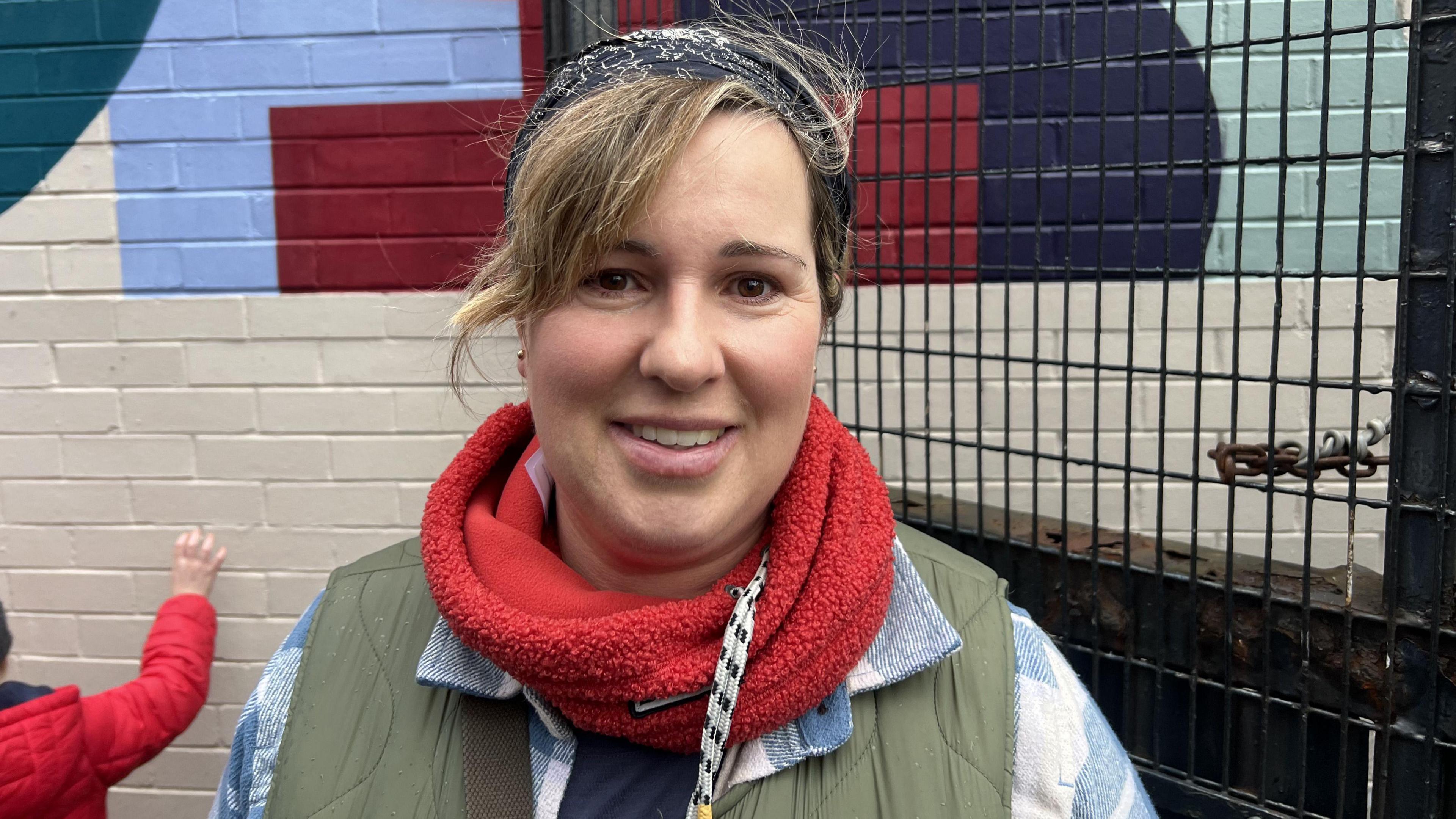 Woman with wearing black headband, a red fluffy snood and a blue and white checked shirt under a green gilet. 