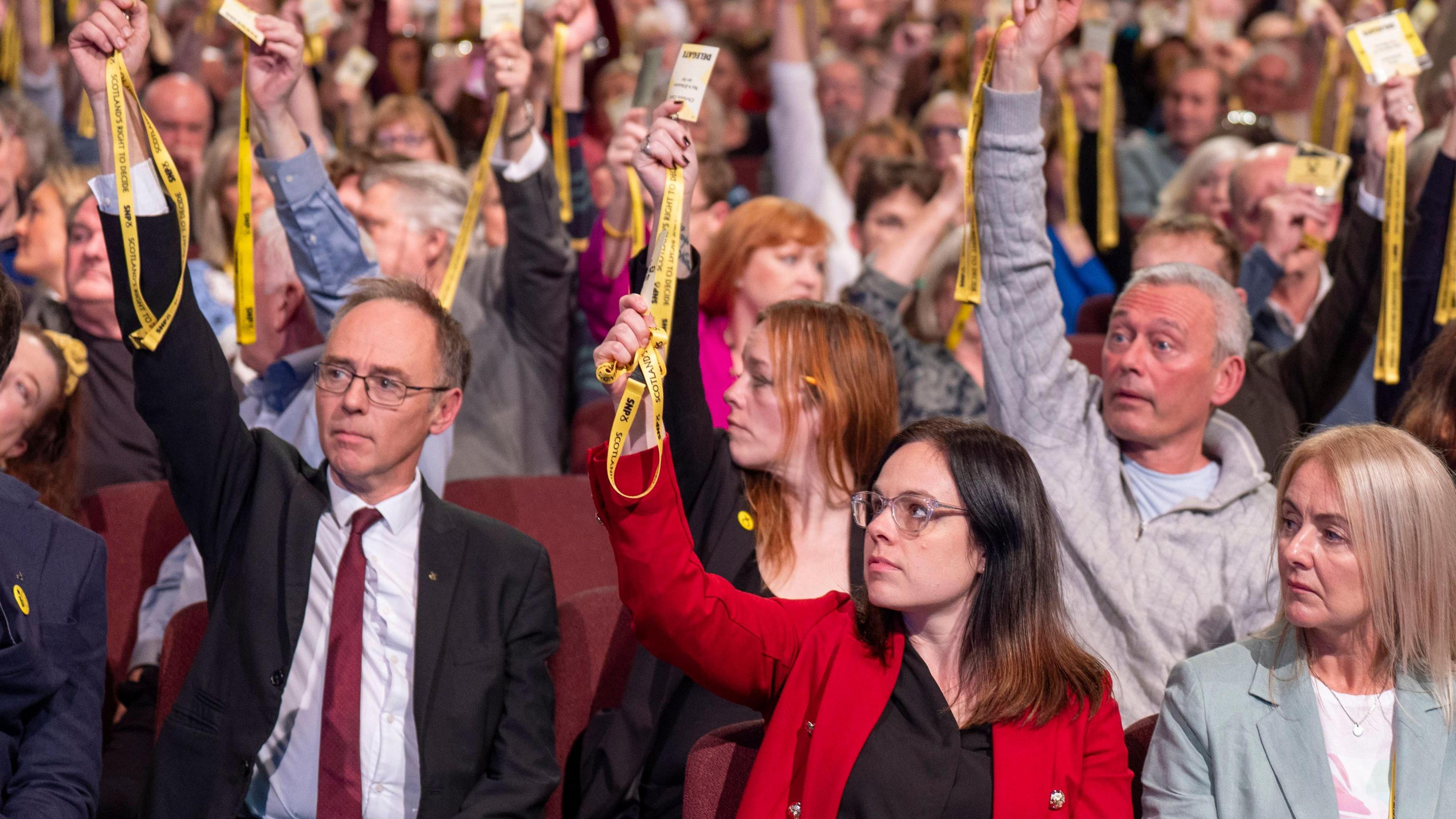SNP members voting on independence strategy. Kate Forbes is in the middle in a red jacket holding a yellow pass.