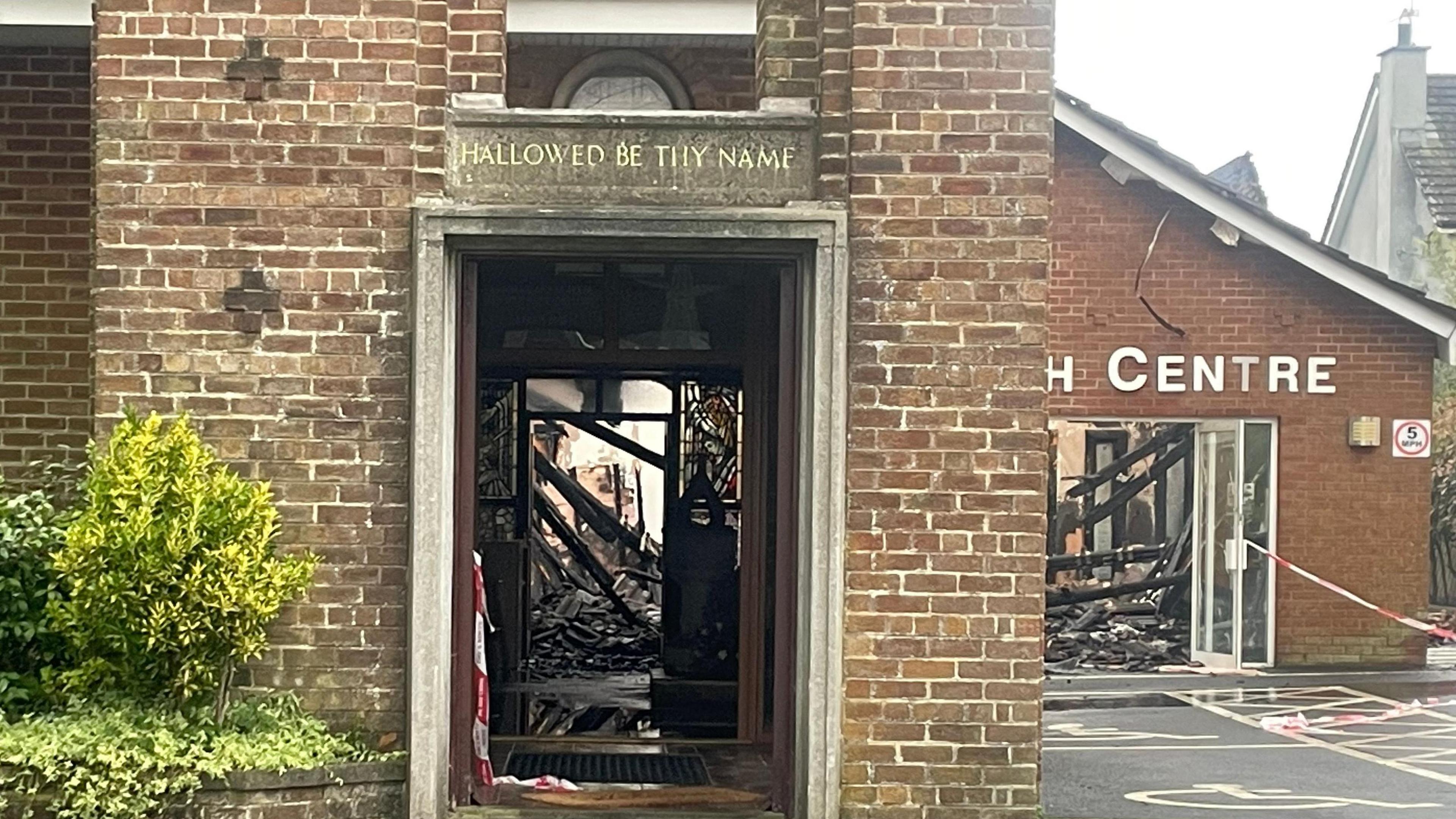 The red brick church and parish centre roof has fallen through and smoke damage is visible through the doors of the buildings.