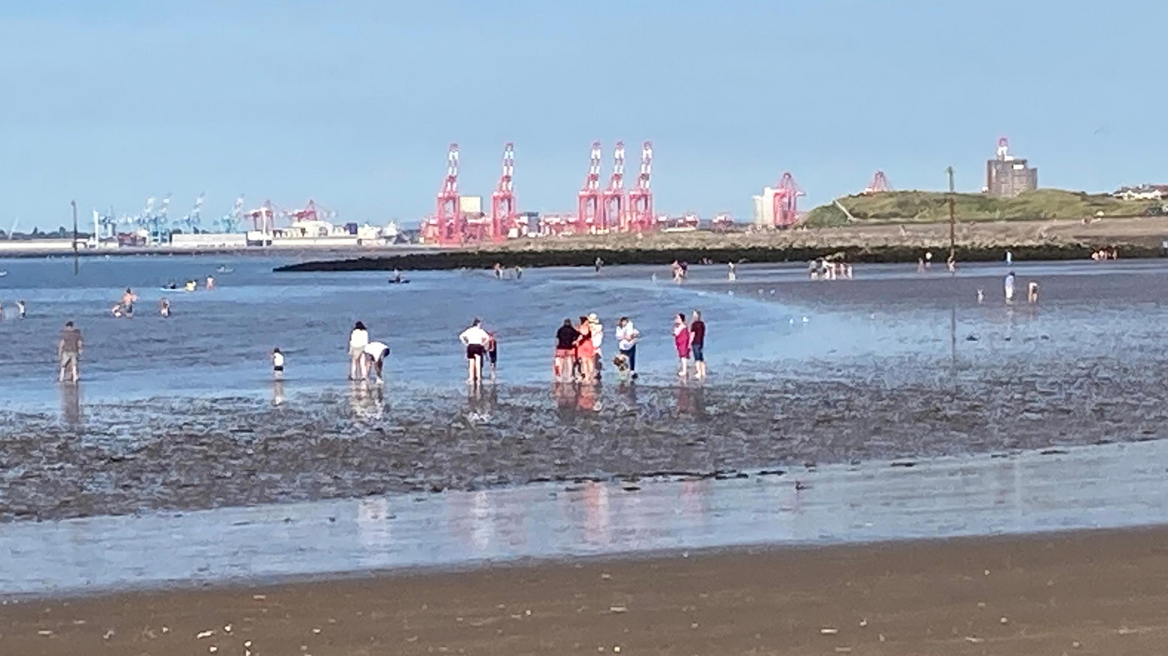 Image shows people on a beach at Leasowe, near New Brighton on the Wirral. There are people paddling in the water. Large red cranes can be seen on the other side of the River Mersey.