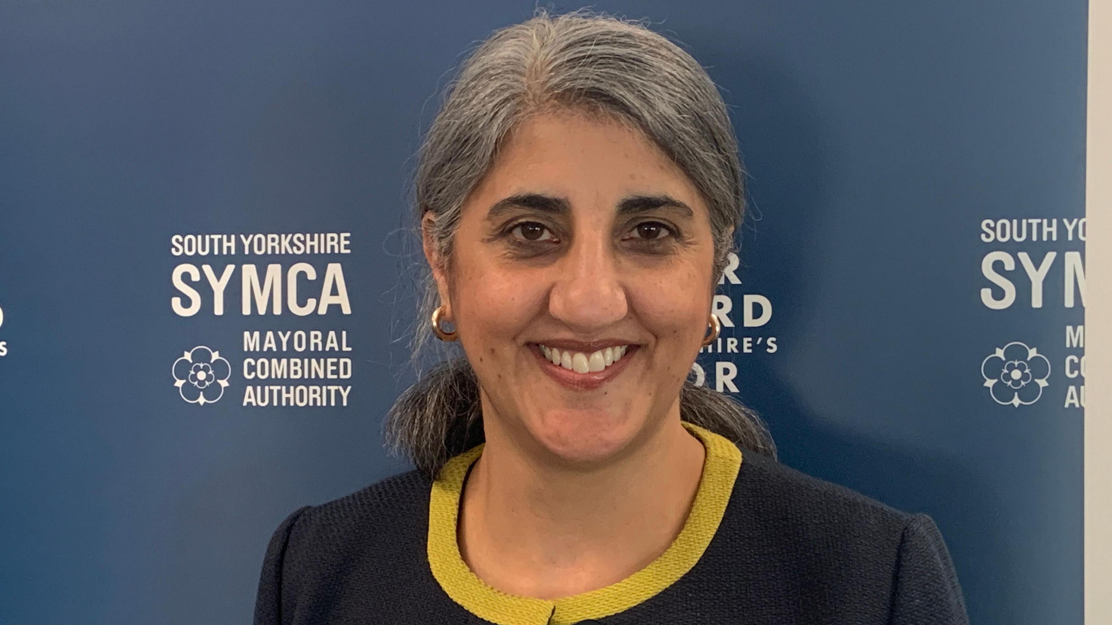 A woman with grey hair is smiling and wearing a navy blue suit with yellow trim. She is stood in front of a blue board which says South Yorkshire SYMCA in white writing