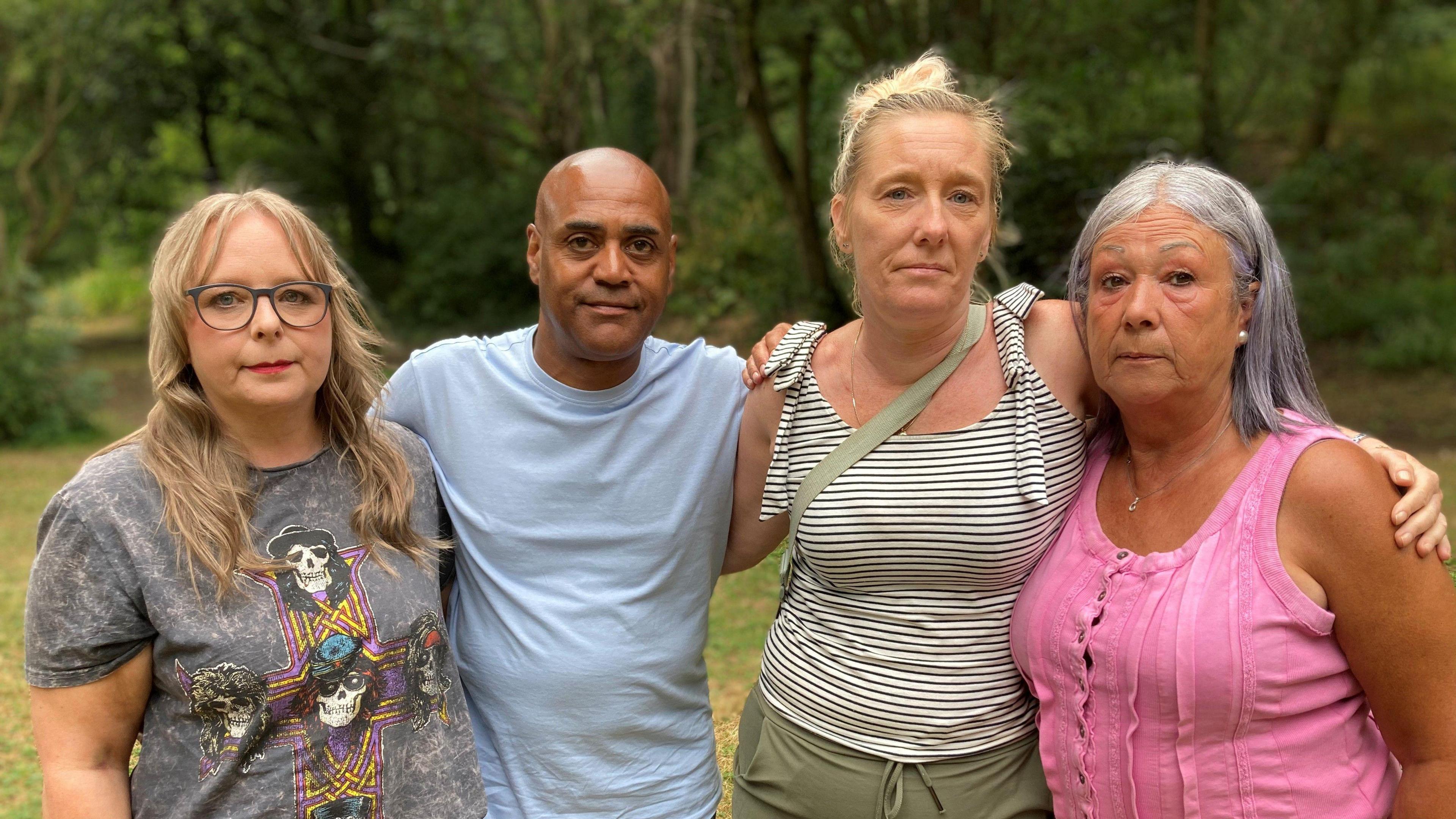 Three women and a man put their arms around each other in an outdoor woodland setting.