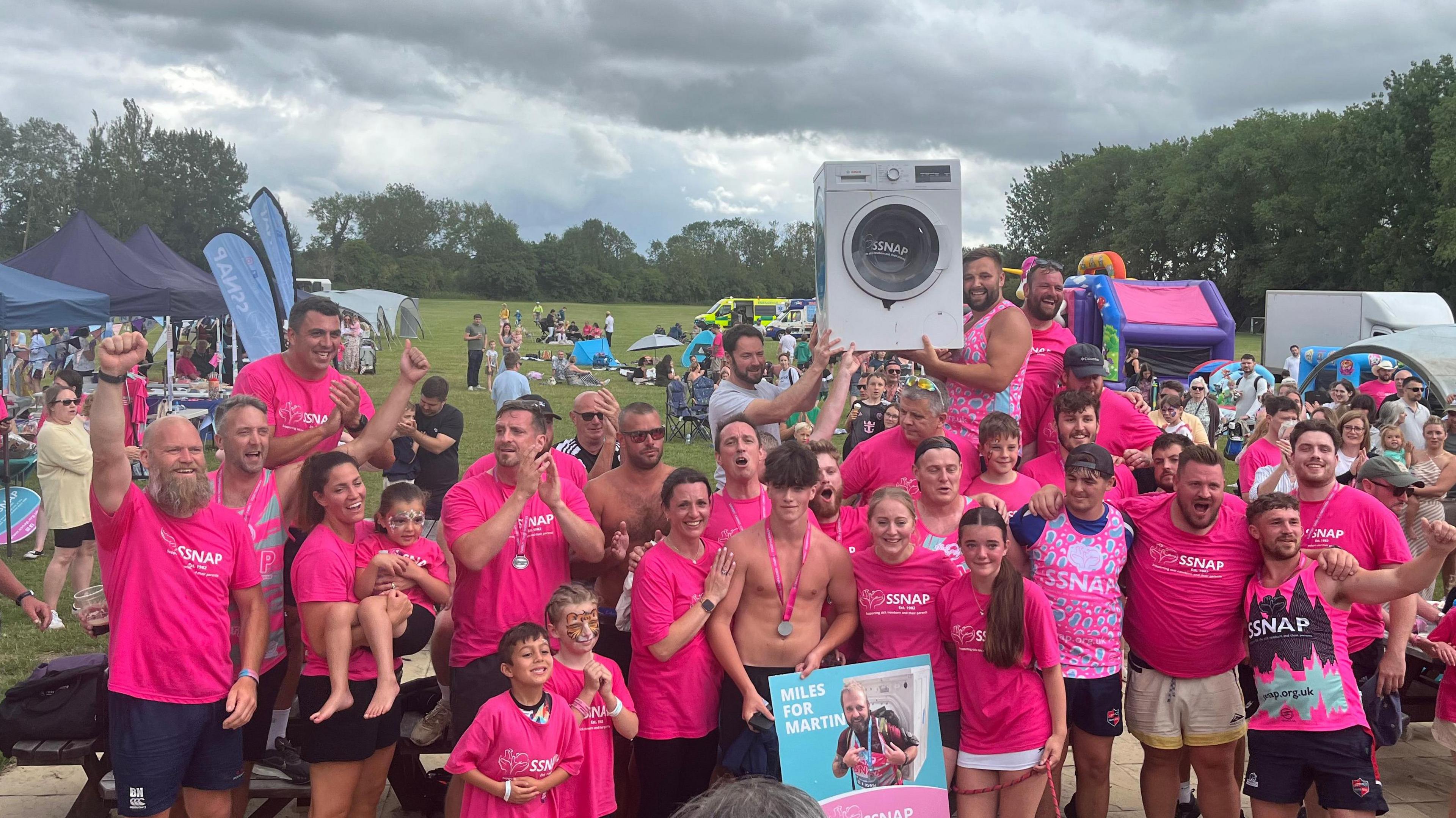In this group photo the runners are joined by partners and children. They are clapping and cheering. The washing machine is held aloft triumphantly.