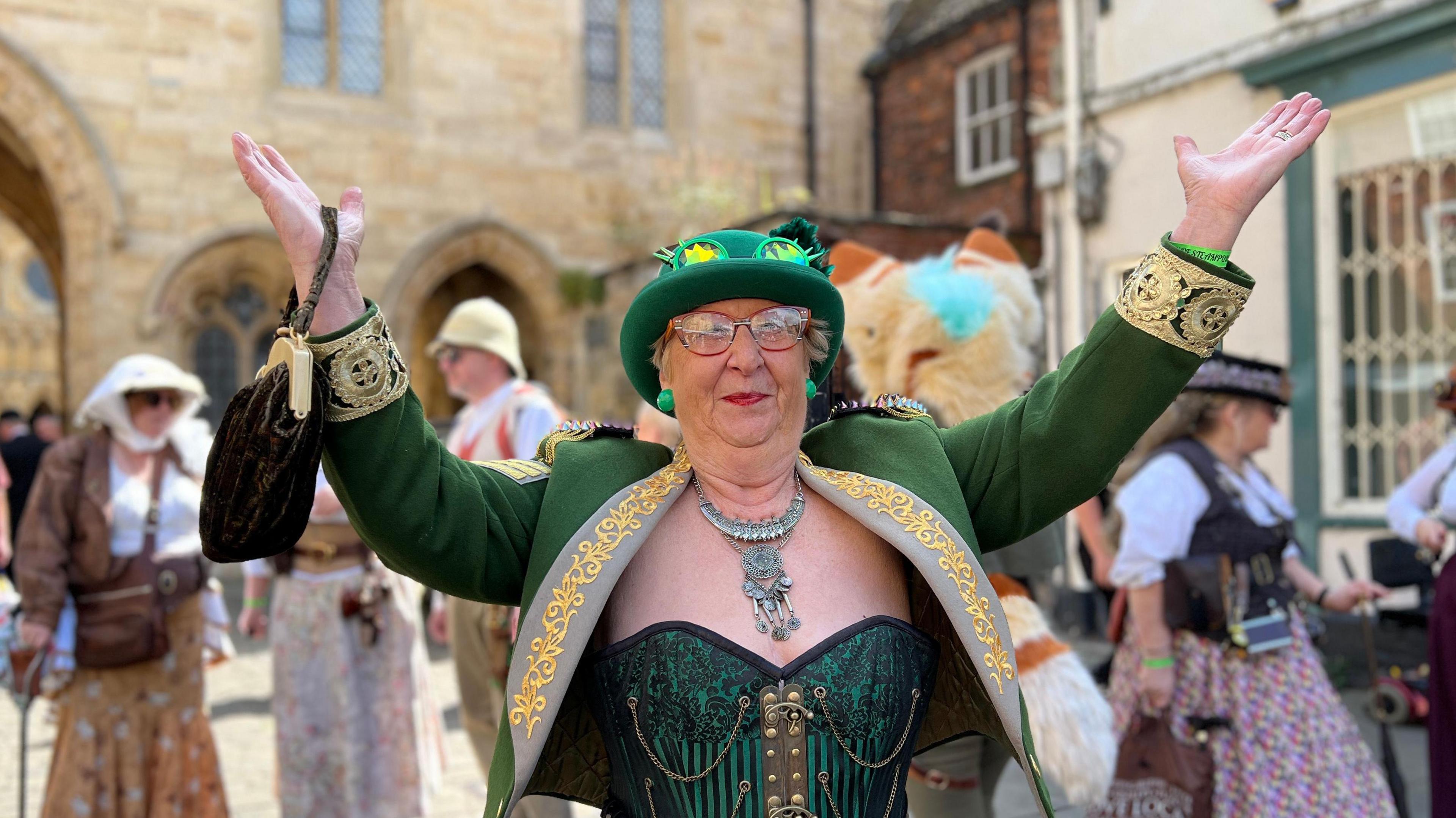 Woman with arms aloft dressed in green top with gold banding on the arms, with a green hat and earrings