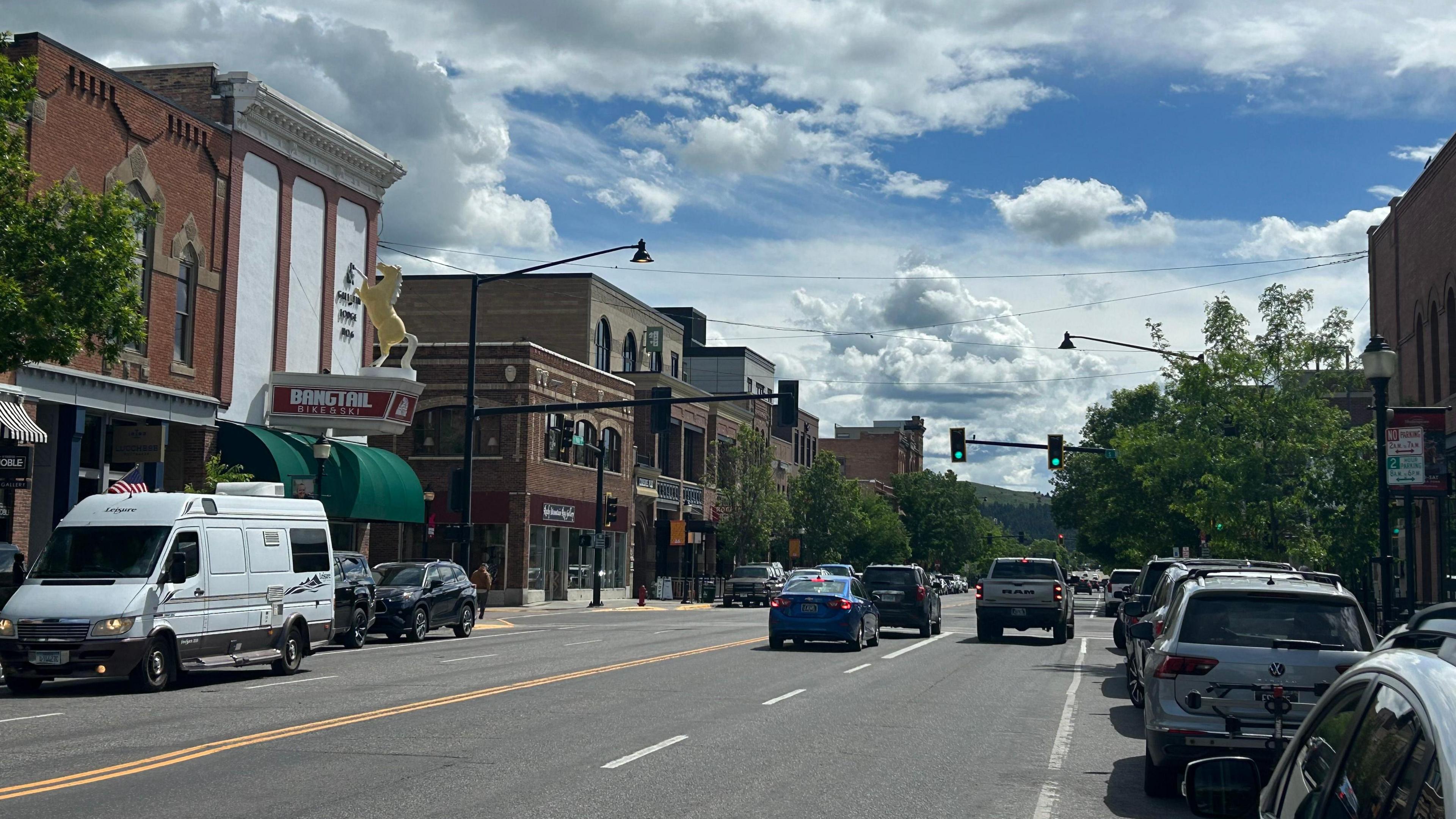 Cars and trucks drive under a blue sky along the main street of Bozeman, lined on either side by parked vehicles and two- or three-storey brick and Western-style buildings housing shops and restaurants