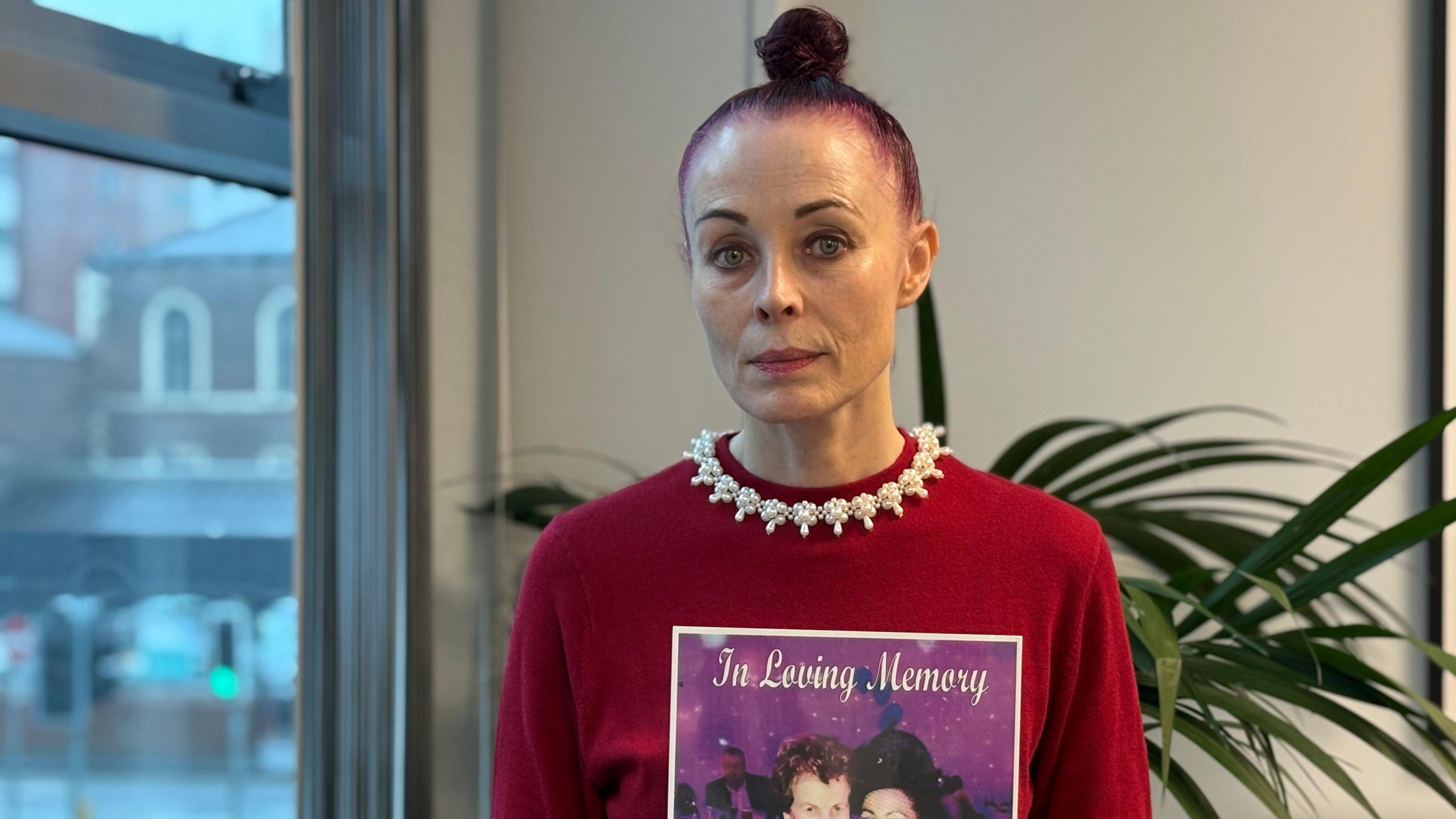 A woman with purple hair looks at the camera as she holds a poster. The poster has a photo of two people, and above it the caption reads 'In Loving Memory'. The woman holding the poster is wearing a red top and a white necklace. 