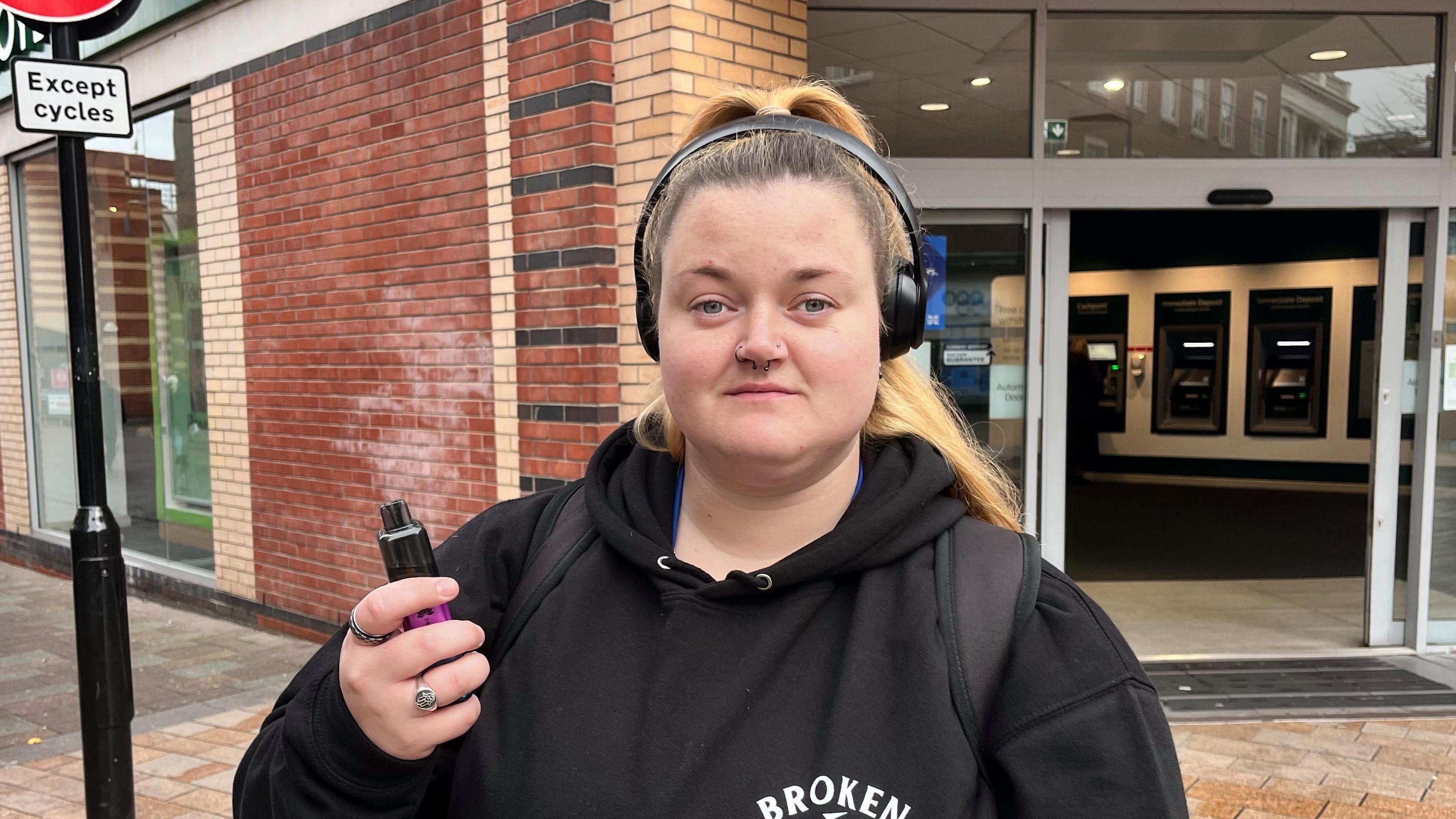 A 26-year-old woman stands in a pedestrian street in front of a high street bank. She is wearing a black hoodie and black headphones and holds a brightly coloured vape in her hand.