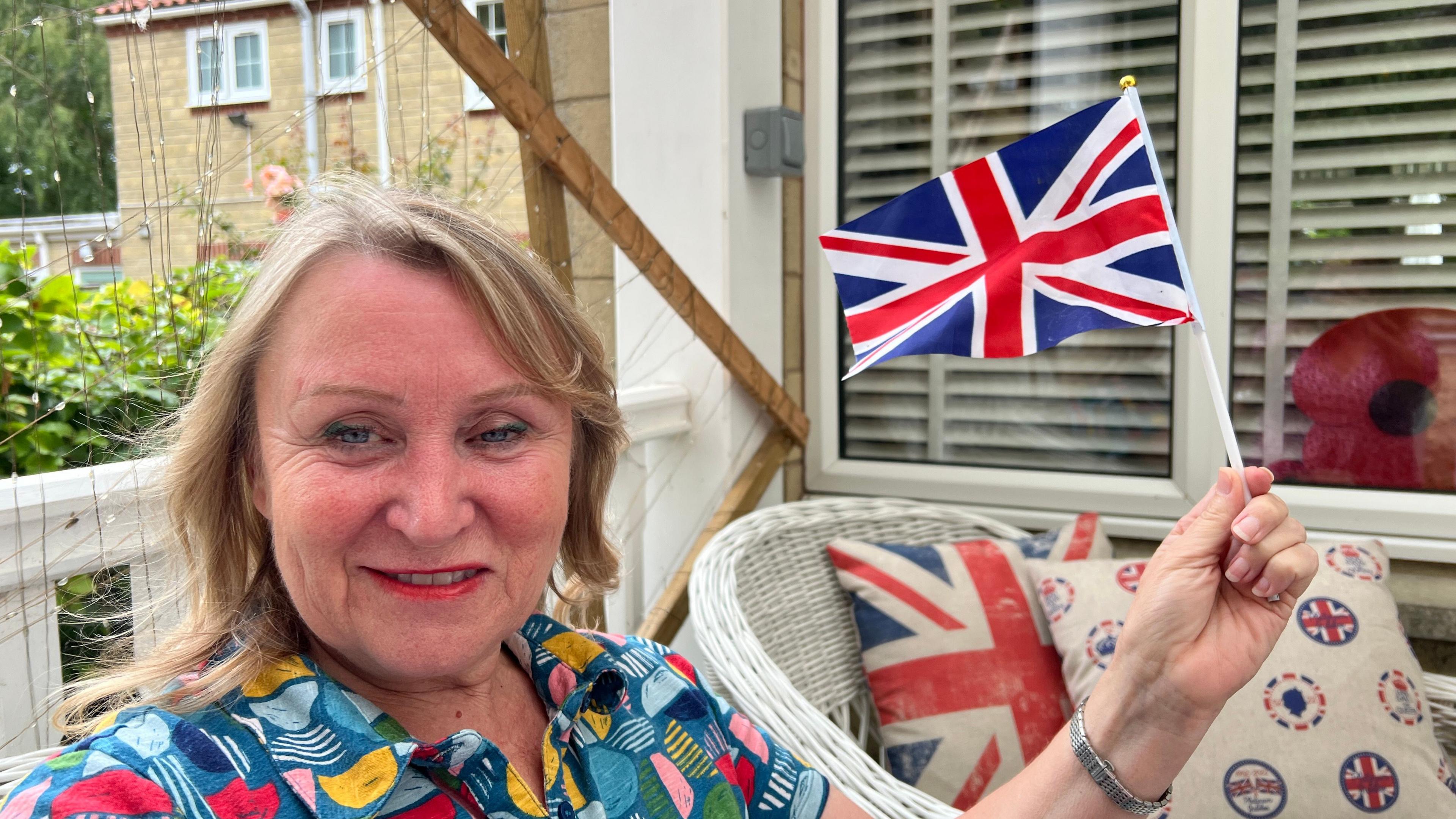 A woman - Kim - wearing a brightly-coloured patterned blouse, holding a union jack flag in her hand. He has short blonde hair and is sat on a patio in a garden. A white wicker chair with union jack cushions can be seen in the background below a window with a cardboard poppy stood against it.