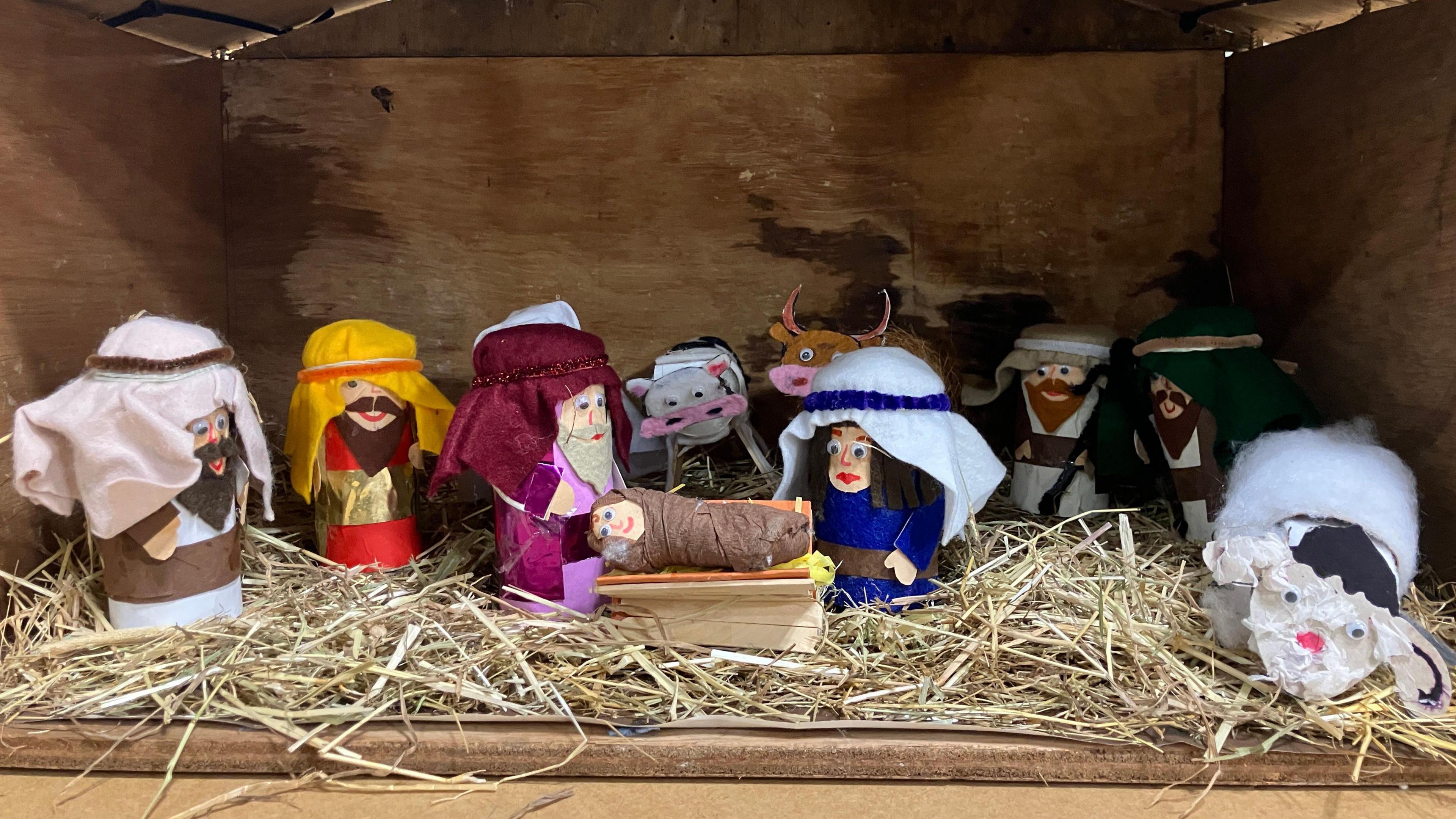 A nativity scene made out of the cardboard tubes from toilet roll. The figures are covered with fabric, pieces of coloured card and pipe cleaners. Joseph, Mary, Jesus and other characters from the nativity are on display in a stable, which contains real straw. 