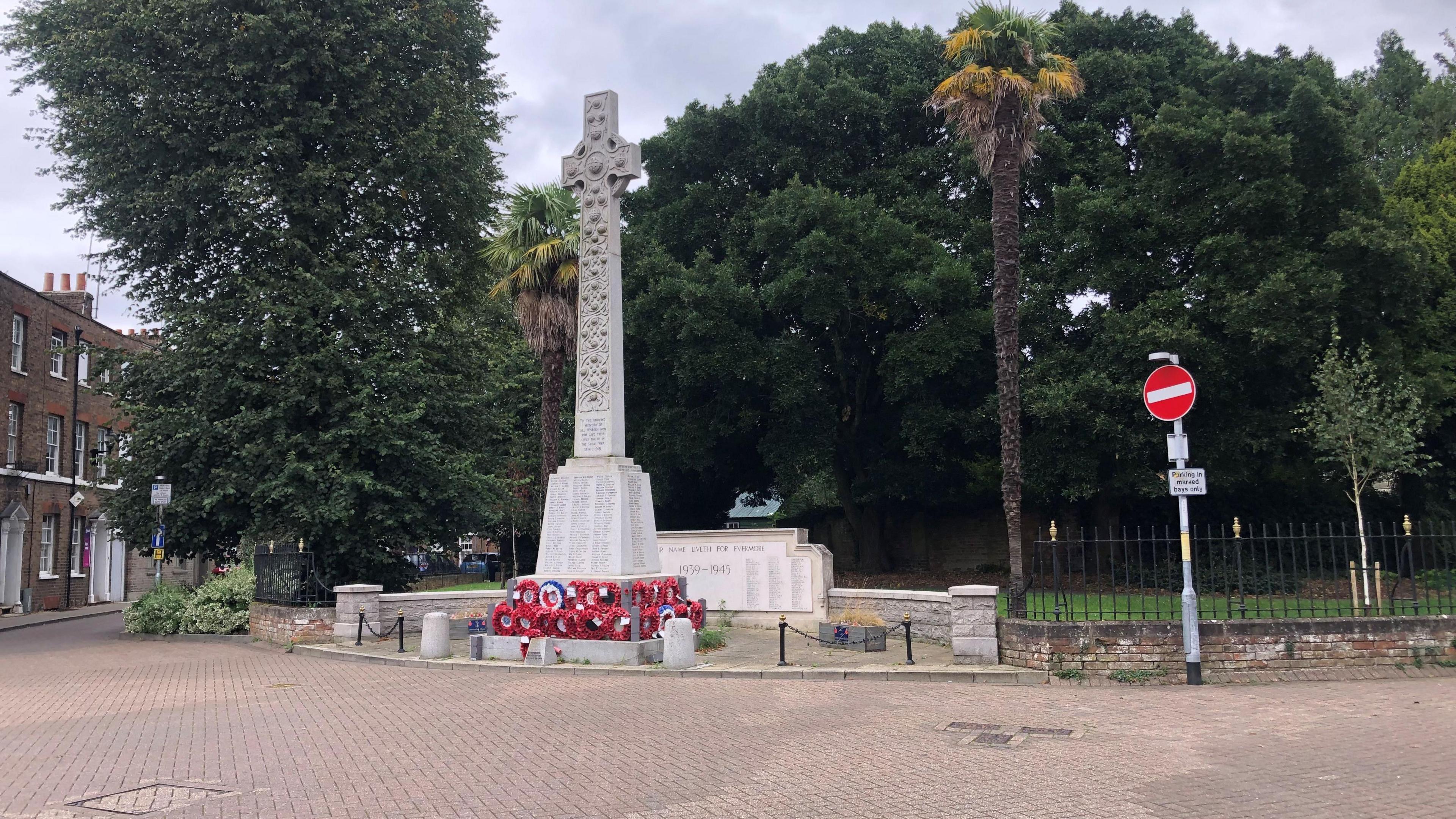 The large stone cross of the war memorial in Wisbech War Memorial Garden. There are poppy wreaths below with a path in front and trees behind with properties in the background.